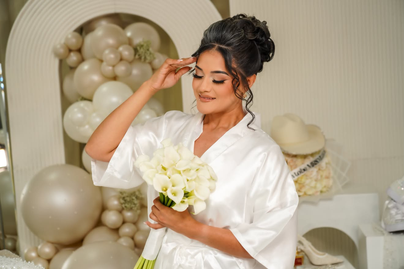Woman in white robe holding a bouquet of white flowers, smiling, with balloons in the background.