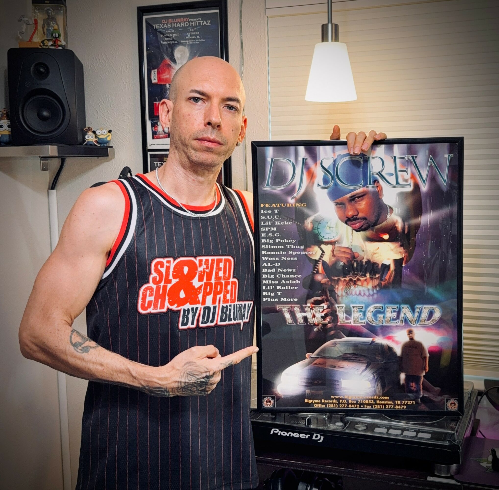 Bald man in a striped basketball jersey points at a framed poster with a car and a person, in a room with a speaker and lamp.