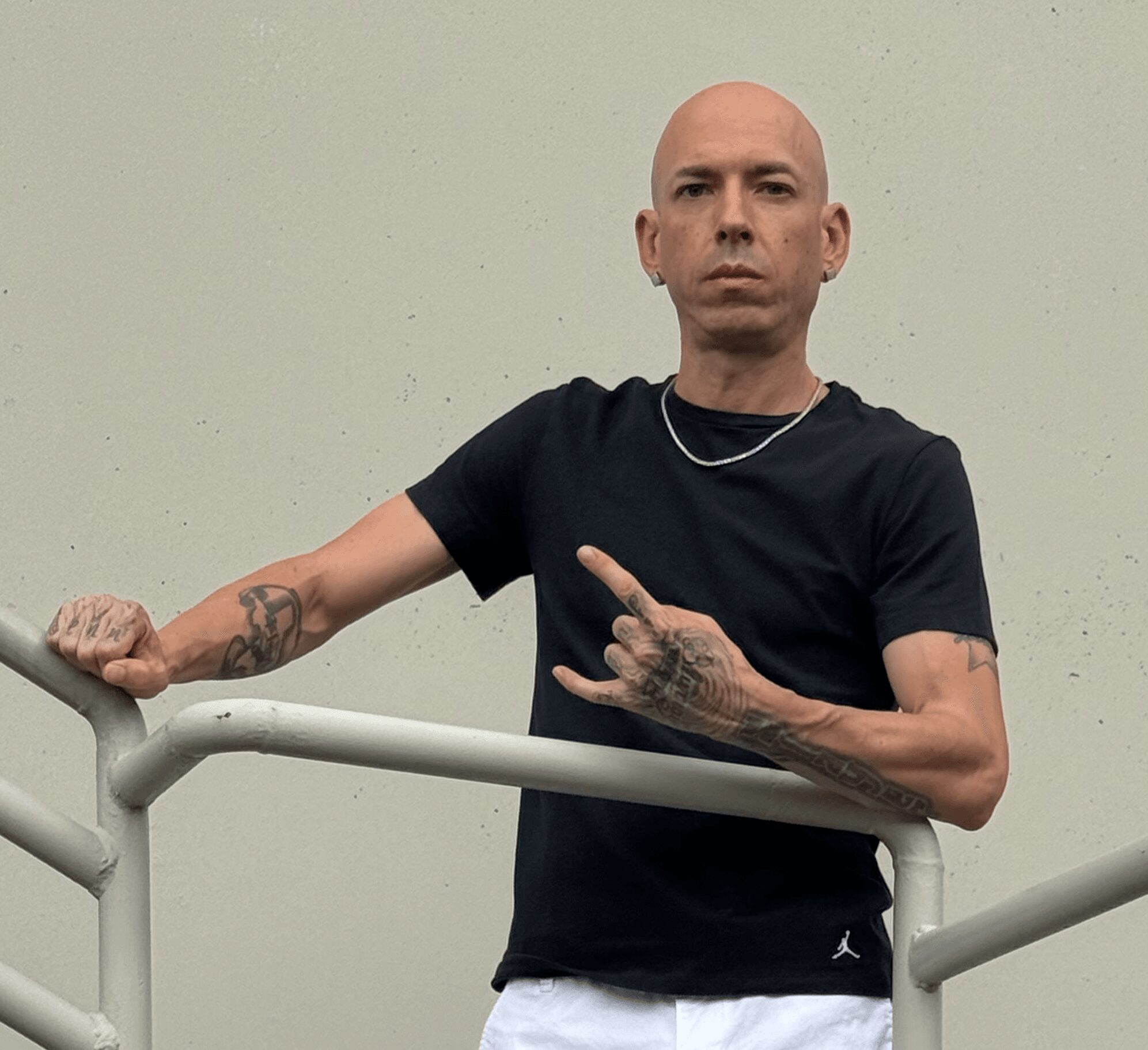 Bald man with tattoos on arms wearing black t-shirt and chain, standing behind a railing against a plain wall.