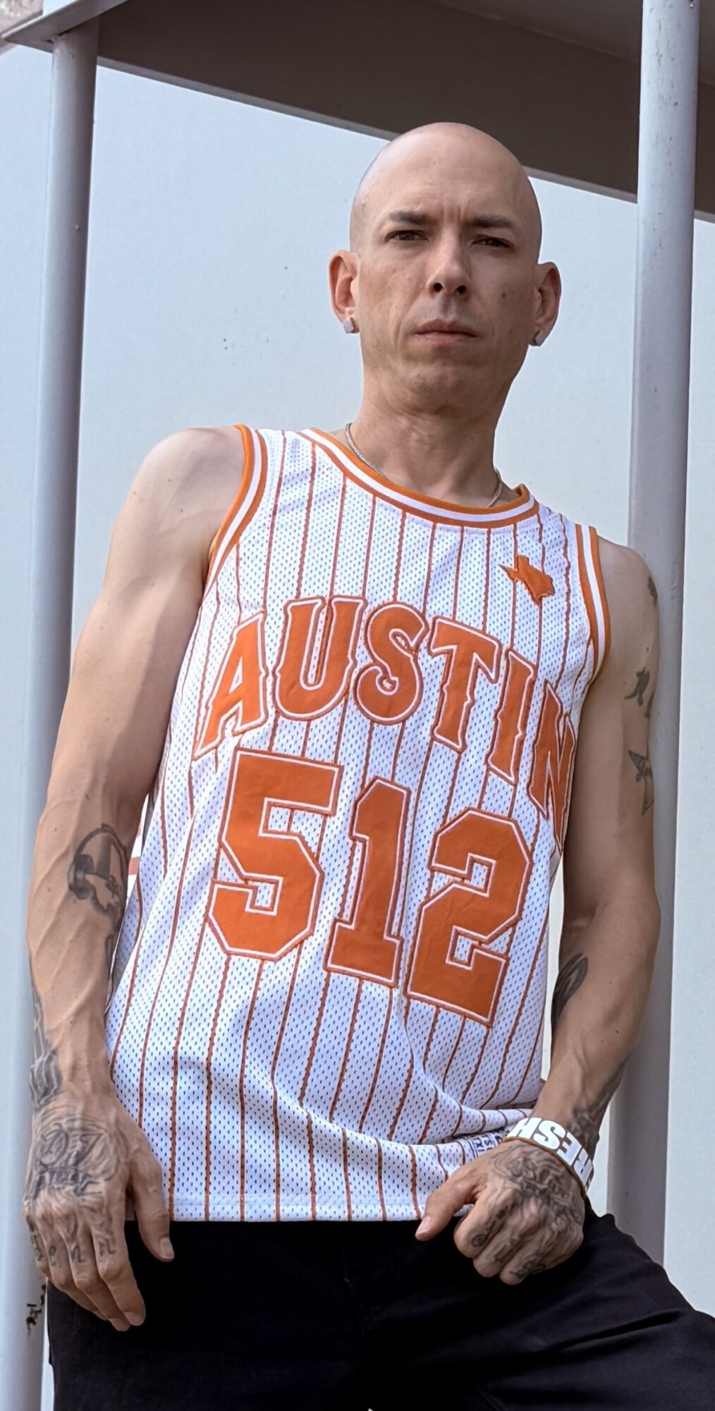 Bald person wearing a striped basketball jersey with 'AUSTIN 512' printed on it, standing outdoors against a blue sky.