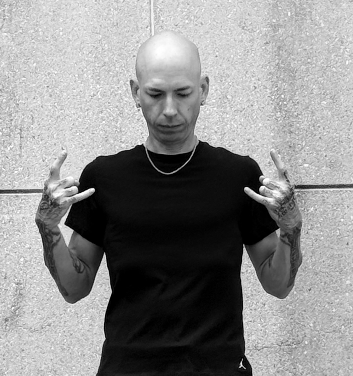 Bald person with tattoos on arms making hand gestures, wearing a black t-shirt and a necklace, standing against a textured wall.
