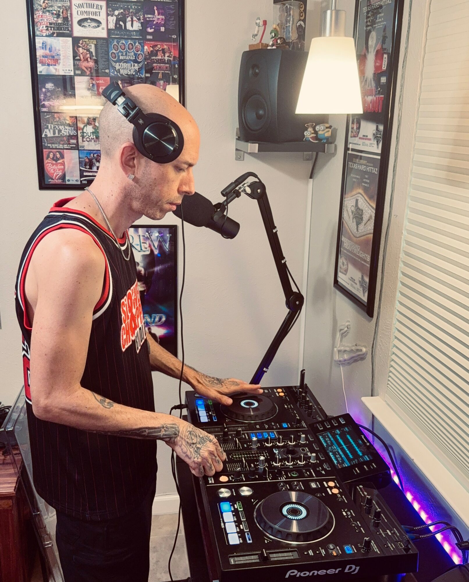 Person with tattoos wearing headphones using DJ equipment in a room with posters and a lamp.