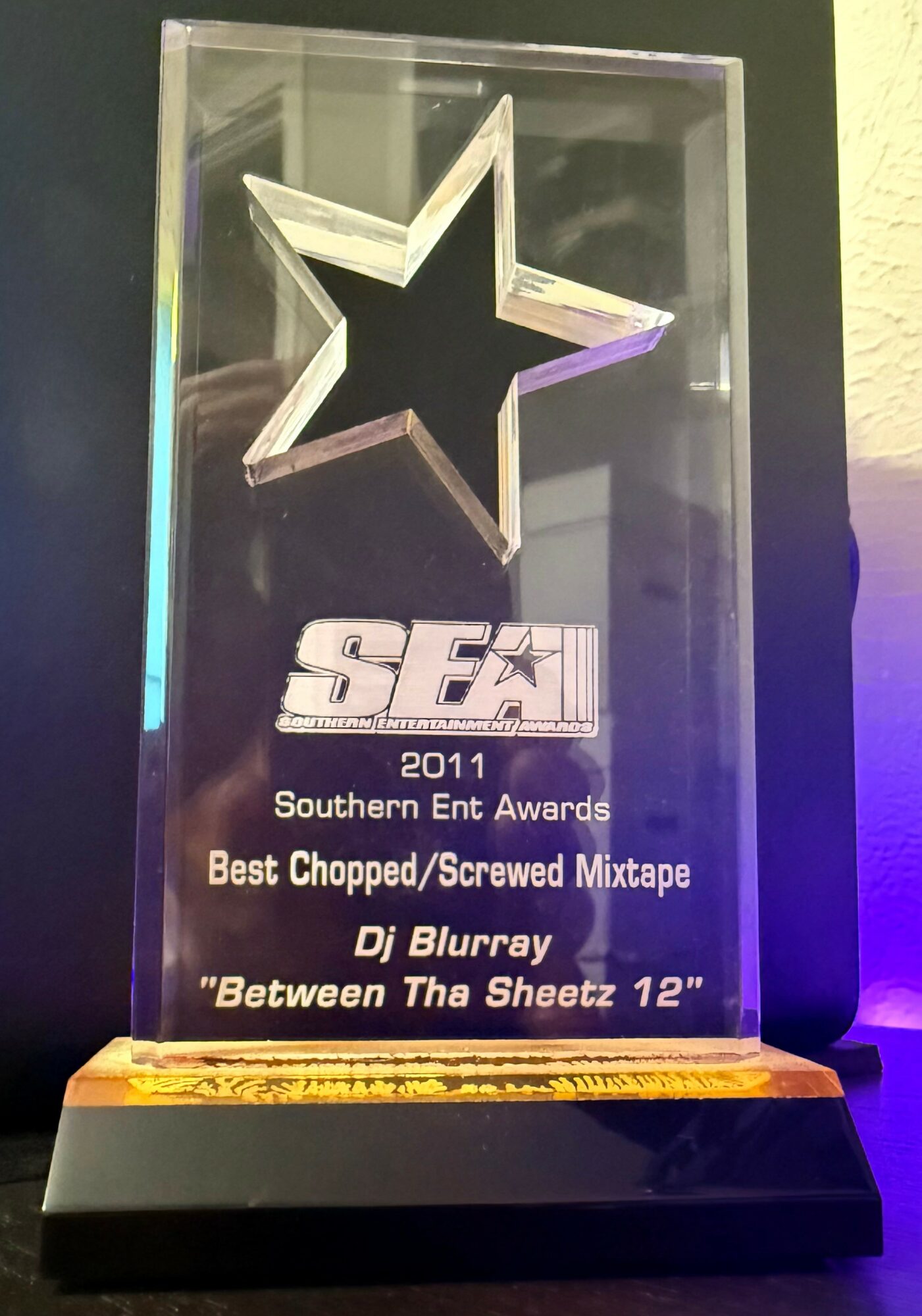 Clear glass award with a star shape, engraved text, and a black base, illuminated by purple and yellow lighting.