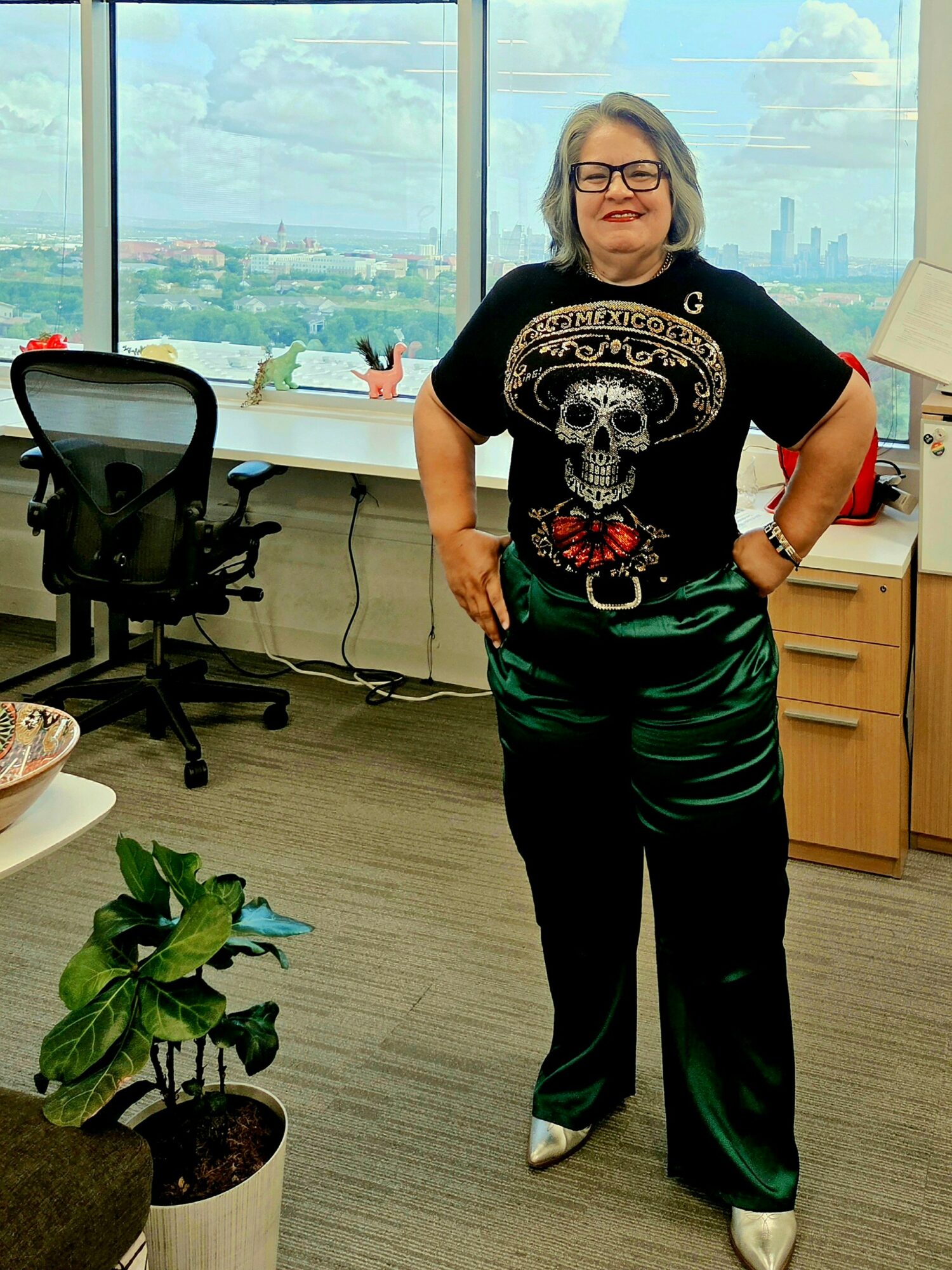 Woman standing in an office with large windows, wearing glasses, a skull graphic T-shirt, and green satin pants, smiling.