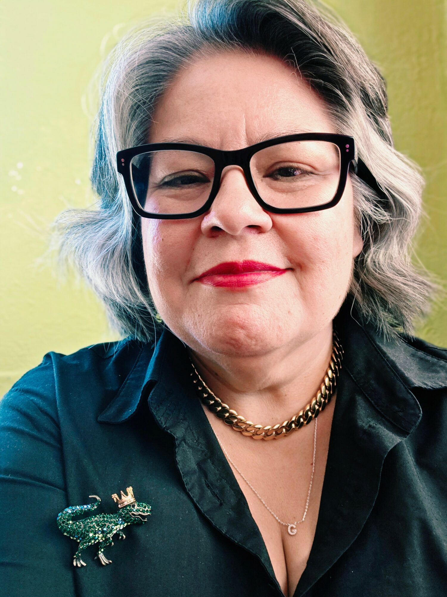 Woman with glasses, gray hair, red lipstick, wearing a black shirt, gold necklace, and a brooch, smiling.