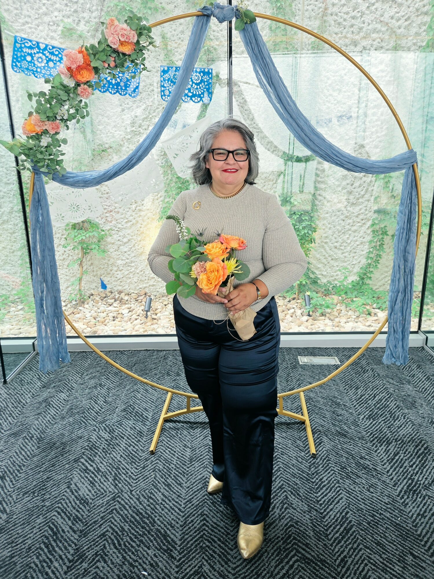 Woman with glasses holding a bouquet of flowers, standing inside a decorative circular frame with blue fabric and flowers, smiling.