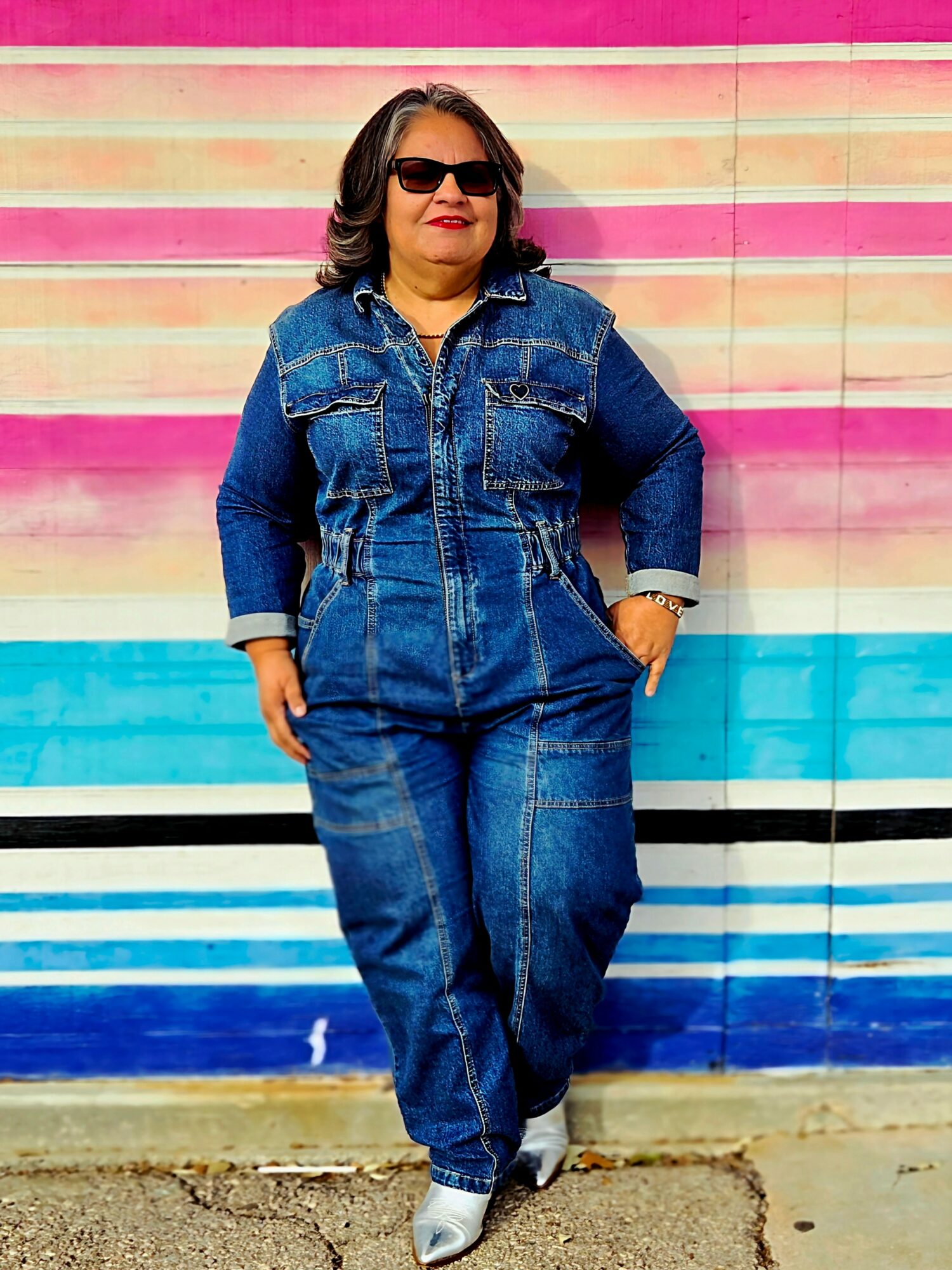 Woman in sunglasses and denim outfit standing against a colorful striped wall, hands in pockets, smiling.