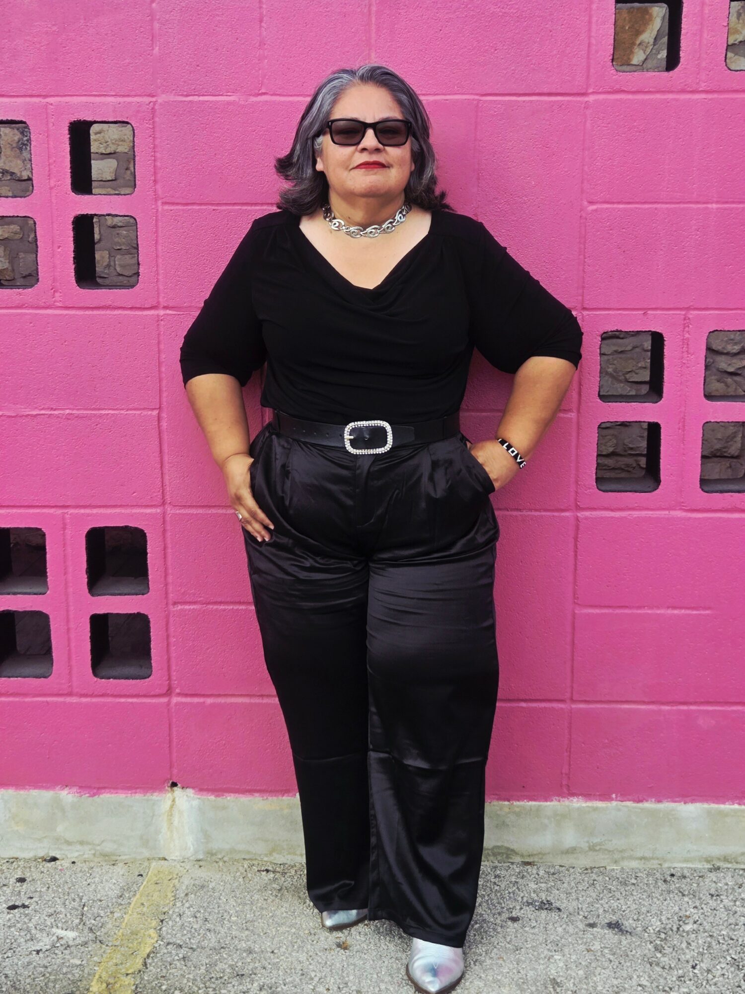 Woman with sunglasses and jewelry stands in front of a pink wall, hands in pockets, smiling.