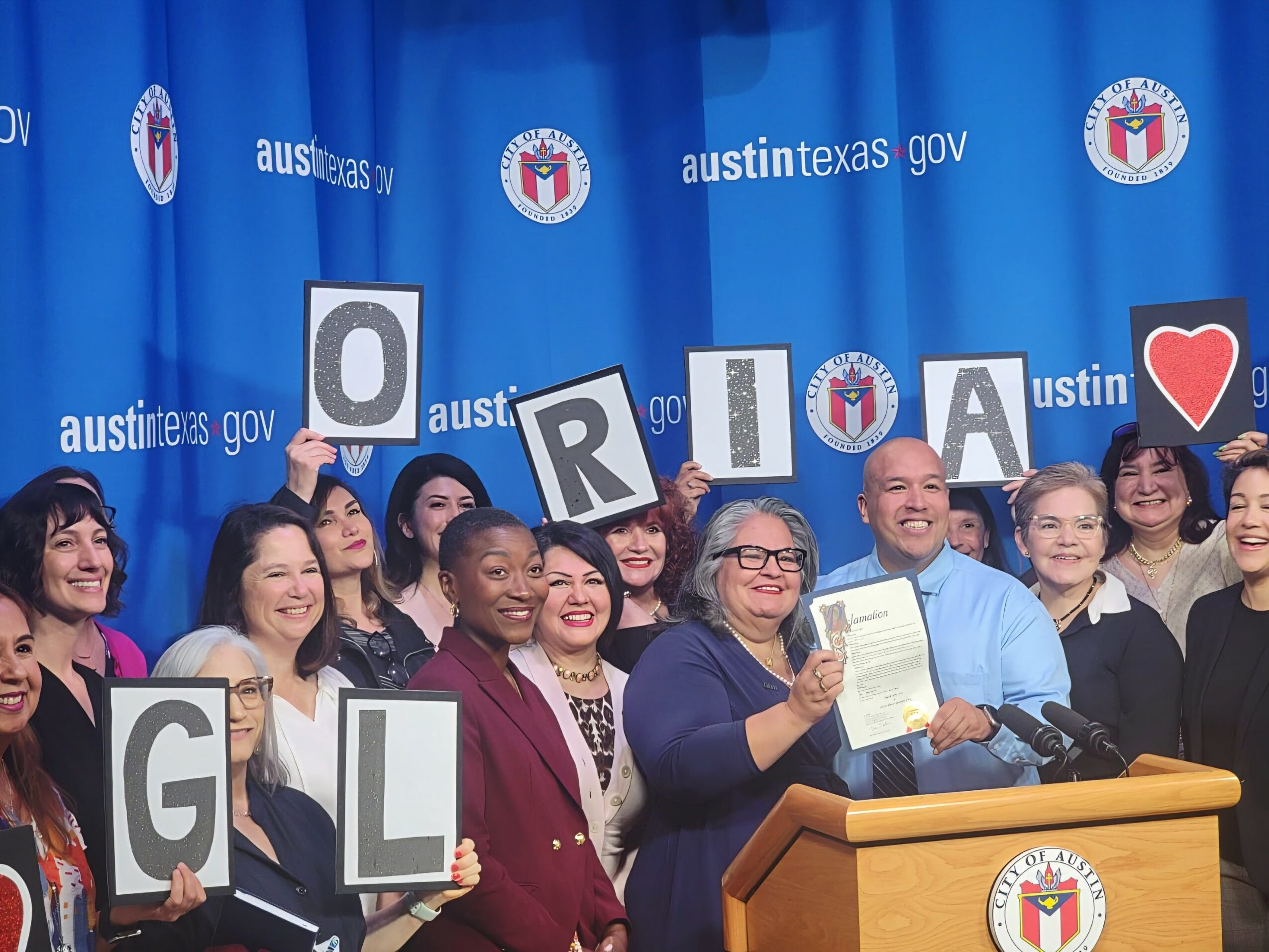 Group of people holding signs spelling 'GUILDER' and a heart, standing behind a podium at an event with a blue backdrop.