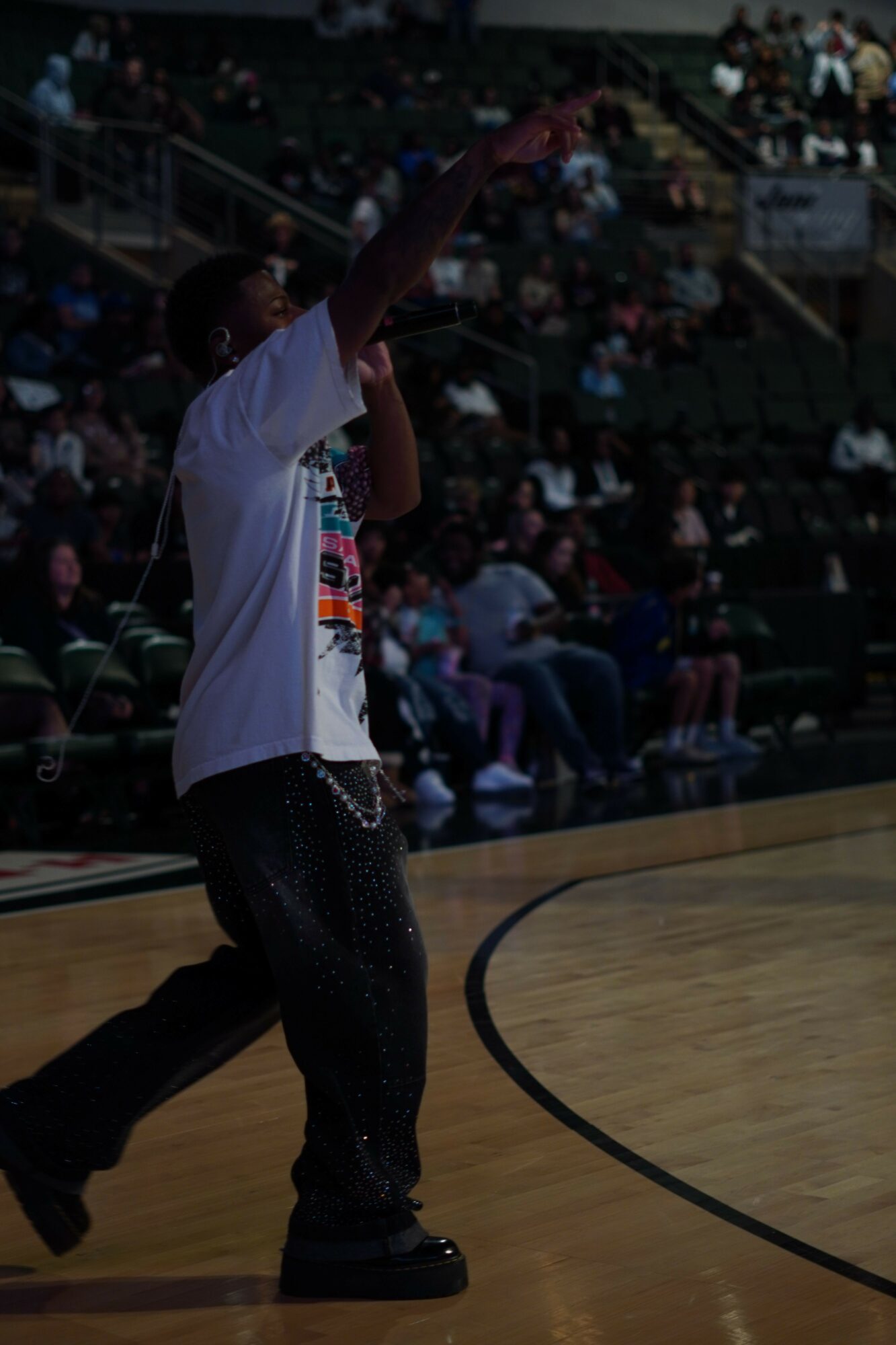 Person standing on a stage with a crowd in the background, arm extended outward, in an indoor setting.