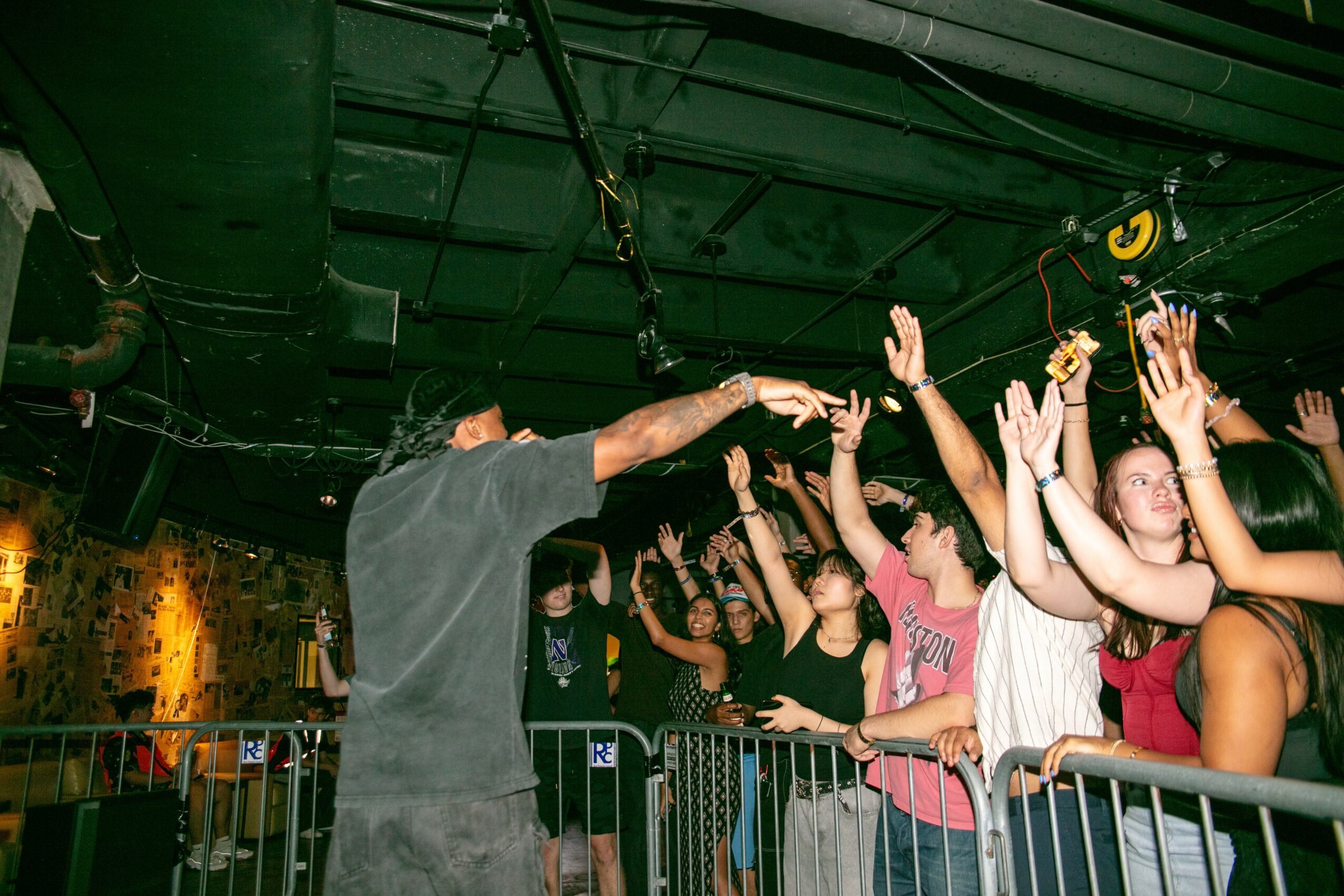 A performer on stage reaches out to audience members with hands raised, in a lively indoor event with crowd behind barriers.