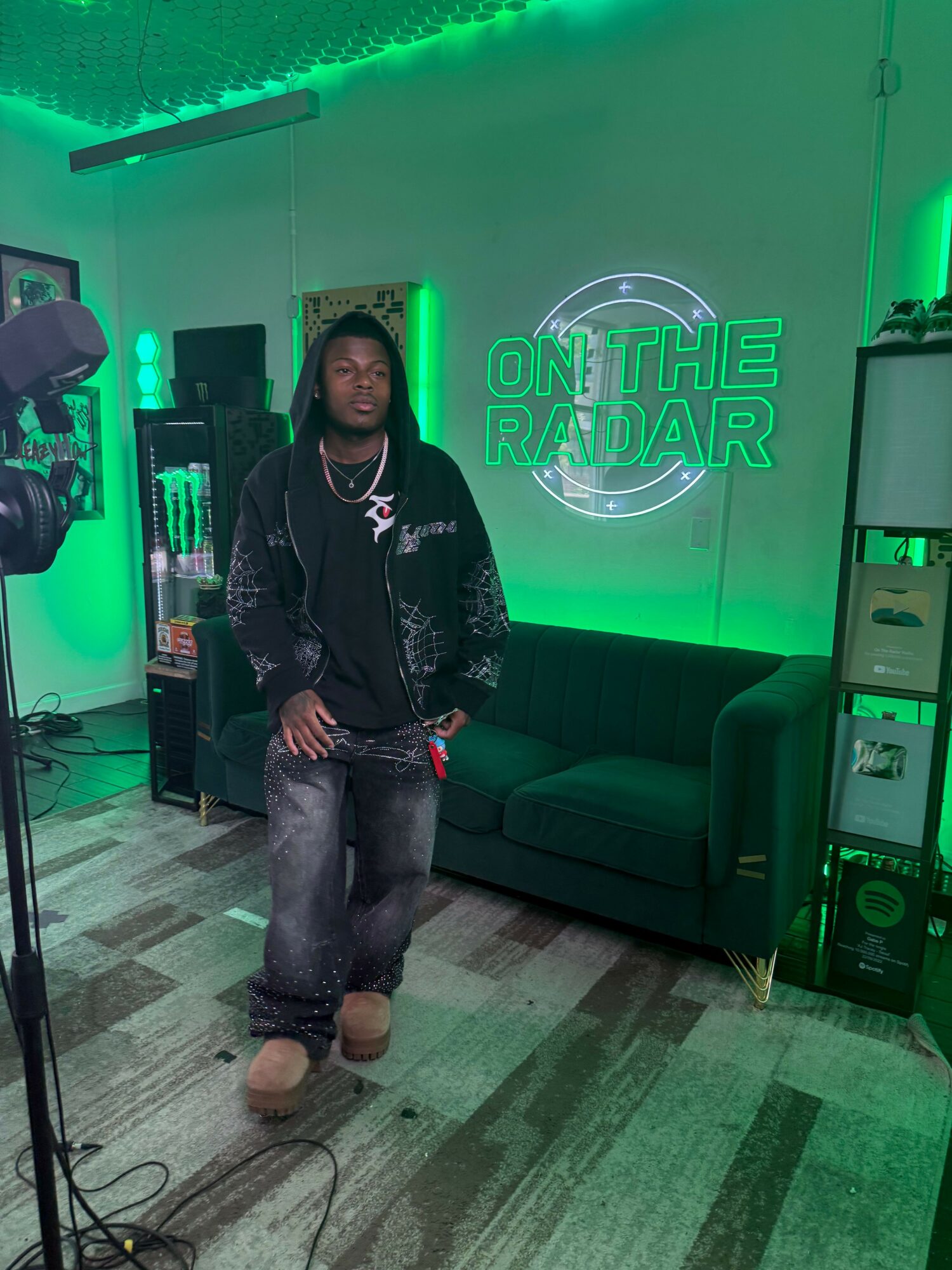 Person standing in a room with green neon lighting, sitting on a green couch, with a sign reading 'ON THE ROCK' behind them.
