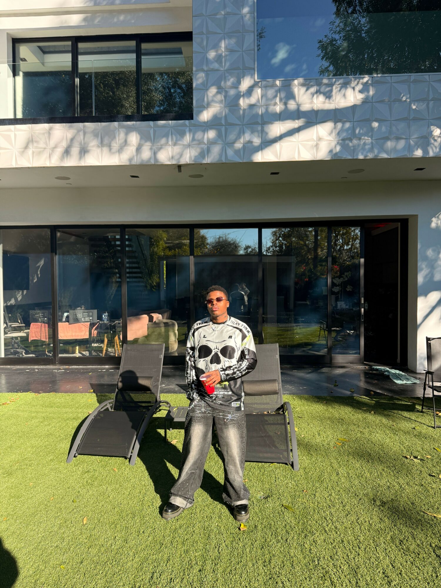 Person sitting on a chair outside a building with glass windows, holding a drink, on a grassy area.