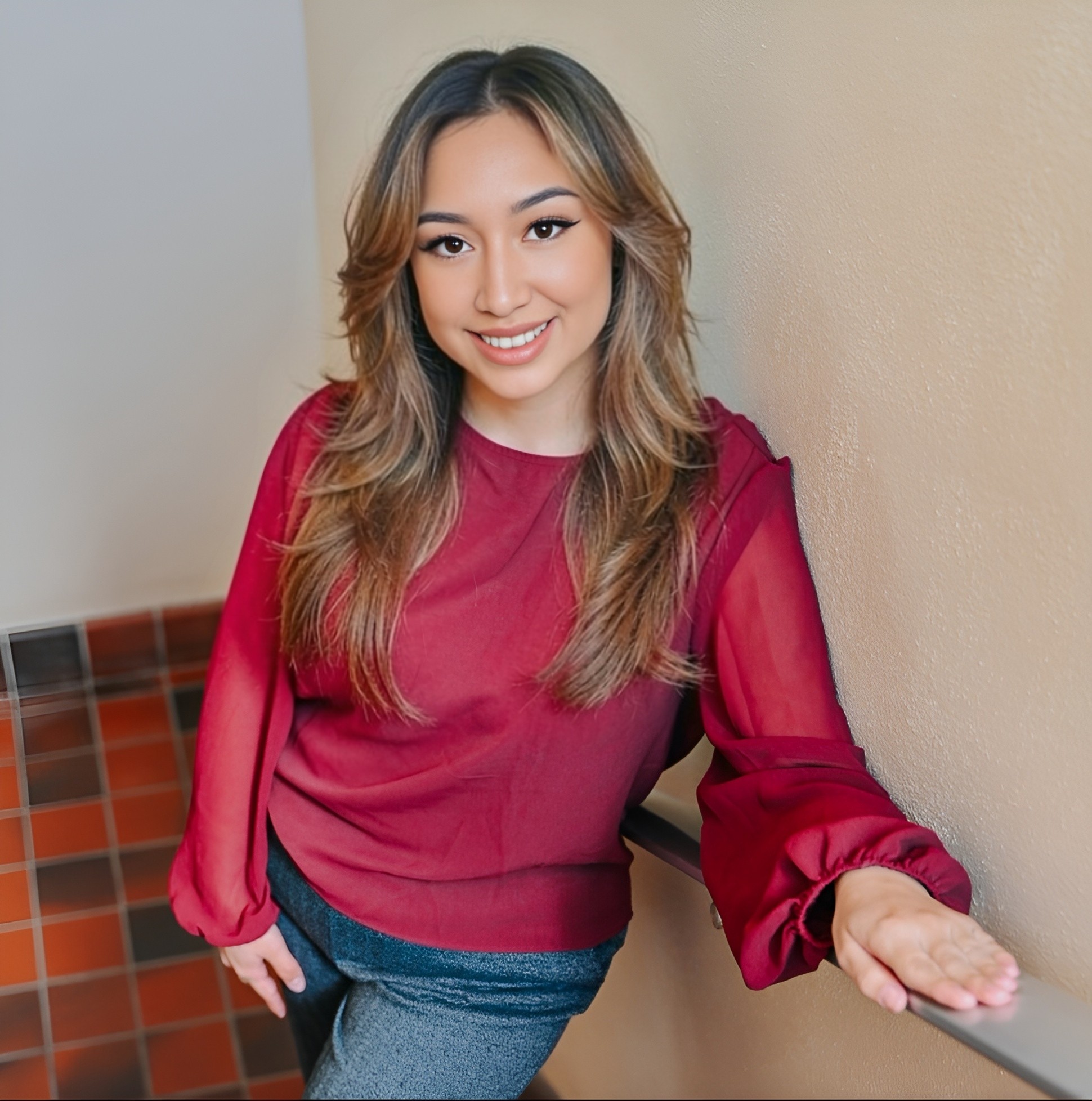 Young woman with long hair smiling, wearing a red blouse, standing against a beige wall, with tiled floor visible.