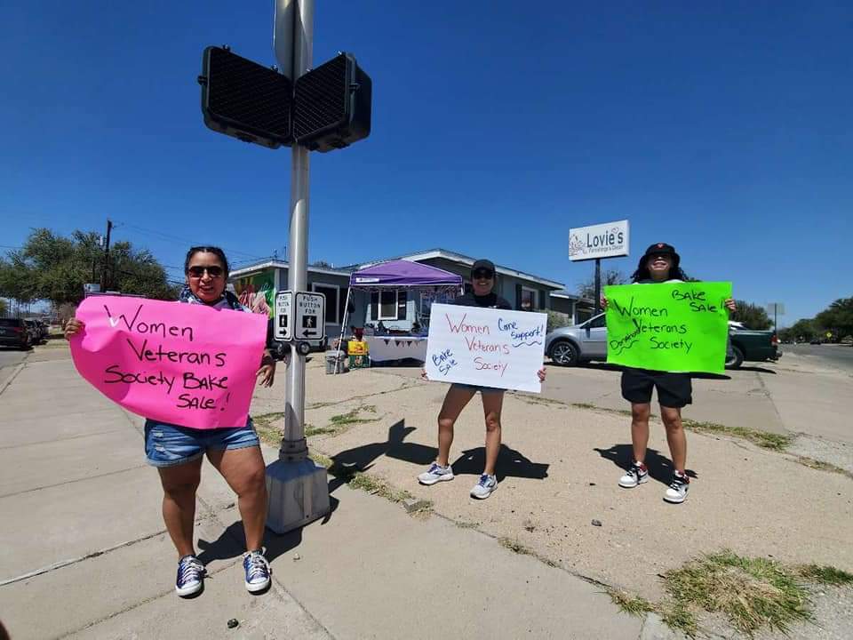 Three women holding signs protest on sidewalk outside a store under a blue sky.