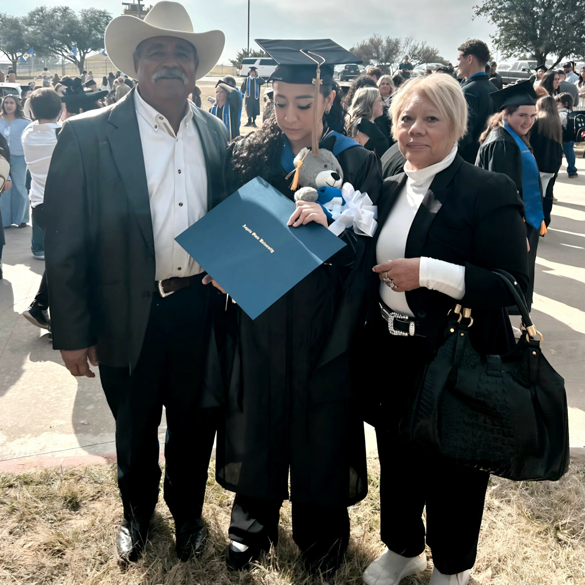 Three people at a graduation ceremony outdoors, one in cap and gown holding a diploma, two others beside them, crowd in background.