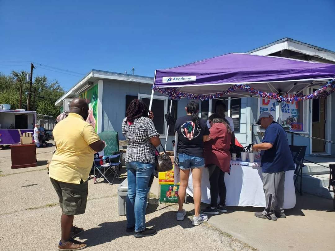 People gather around a table under a purple canopy outside small buildings on a sunny day.