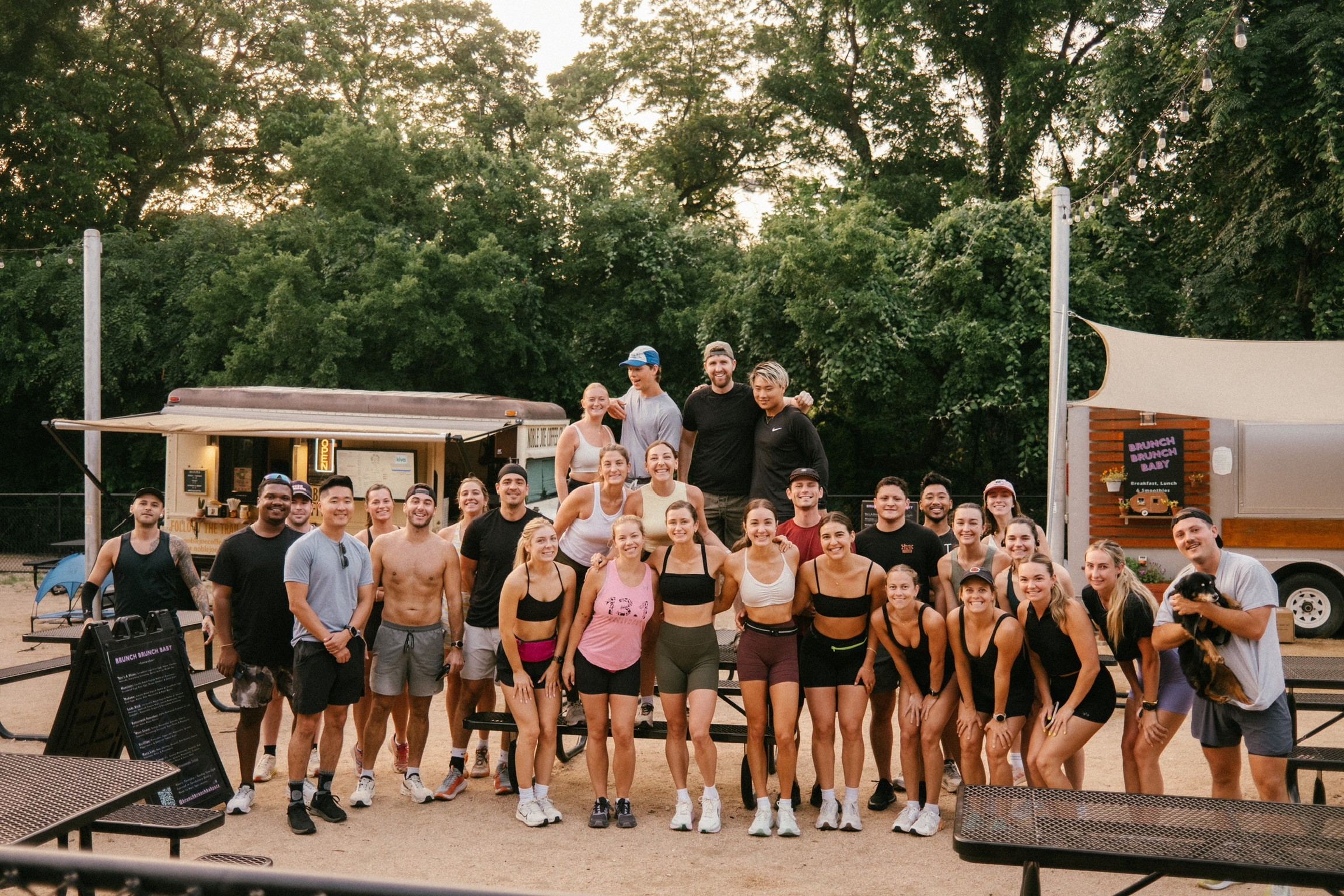 Group of people posing outdoors in front of trees and a building, some wearing athletic clothing, smiling.