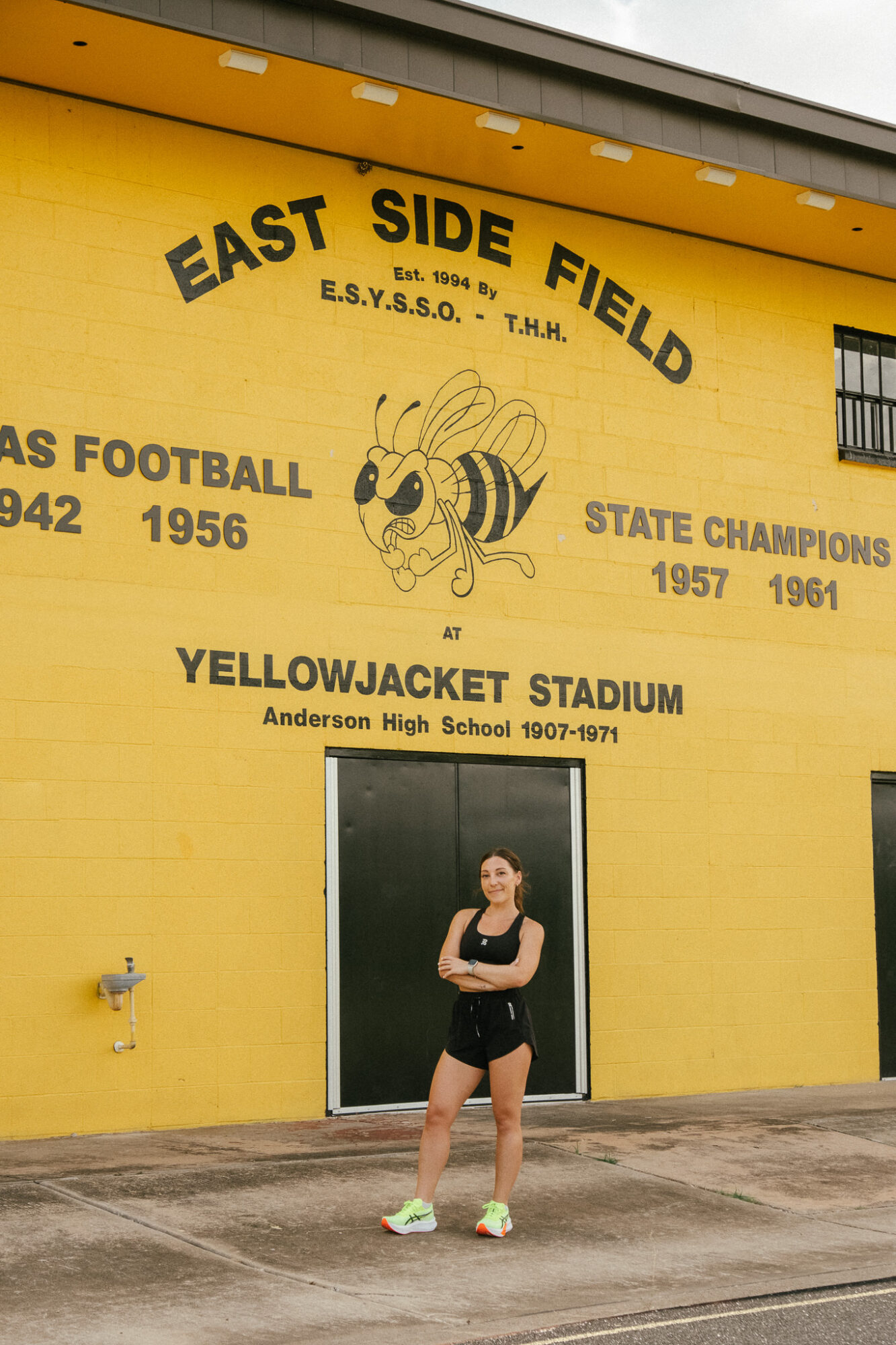 Person standing in front of a yellow wall with a bee mascot and text about East Side Field and Yellowjacket Stadium, with a door below.