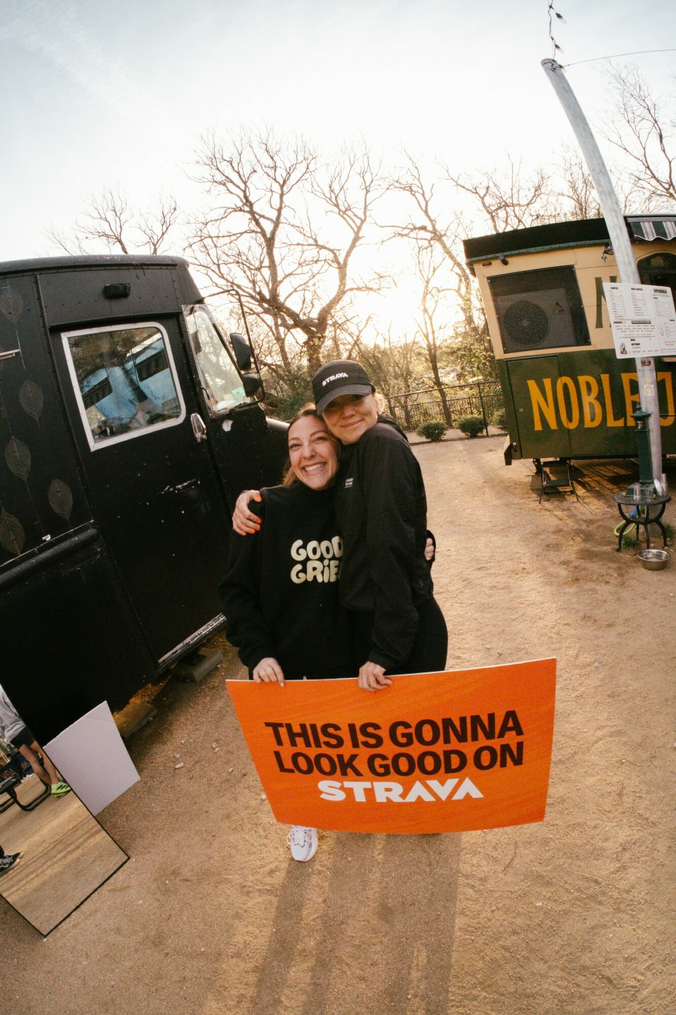 Two people smiling holding a bright orange sign outdoors, trees and a black vehicle in the background.