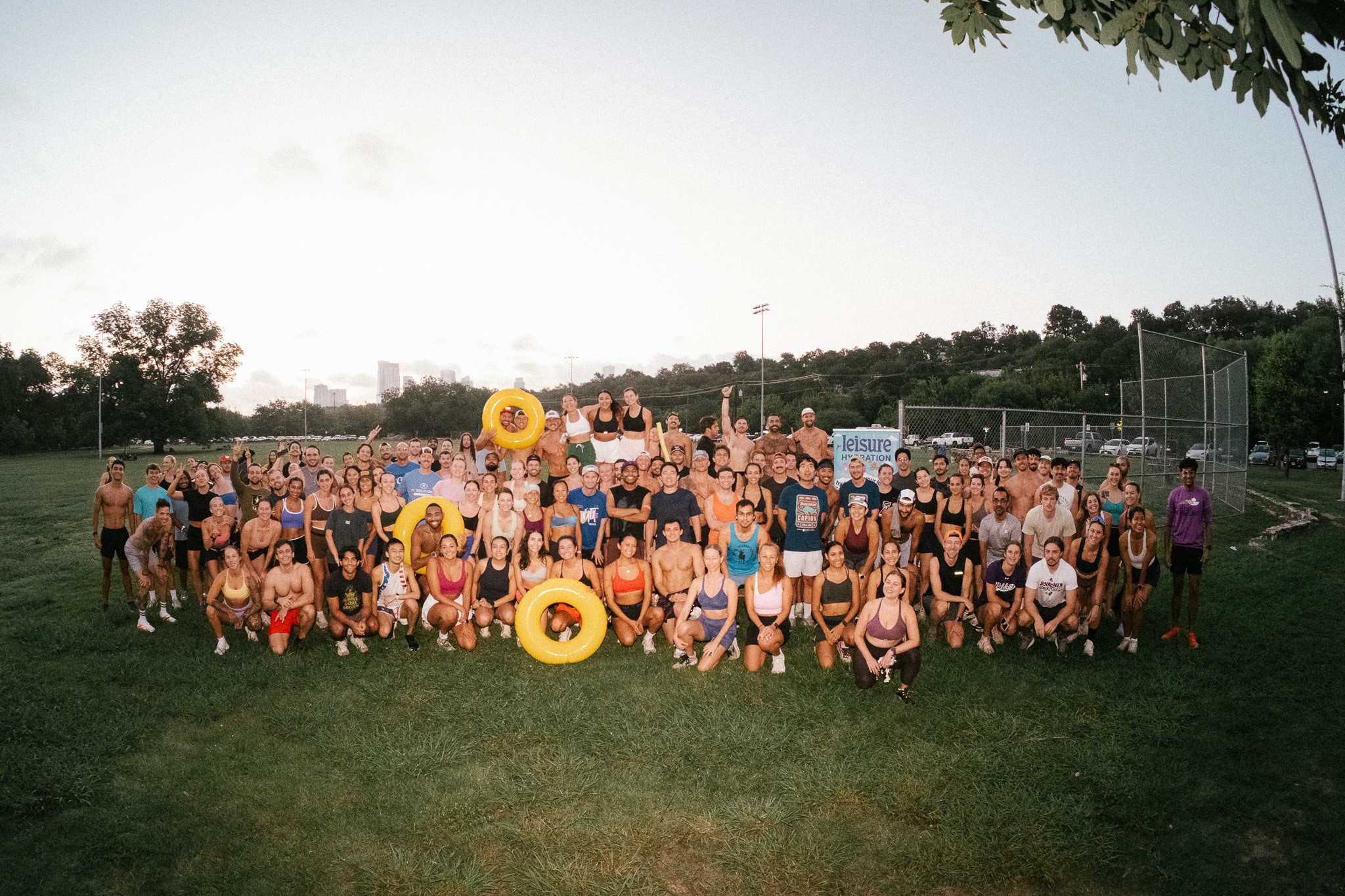 Large group of people gathered outdoors on grass, with trees and a sports field in the background, under a clear sky.