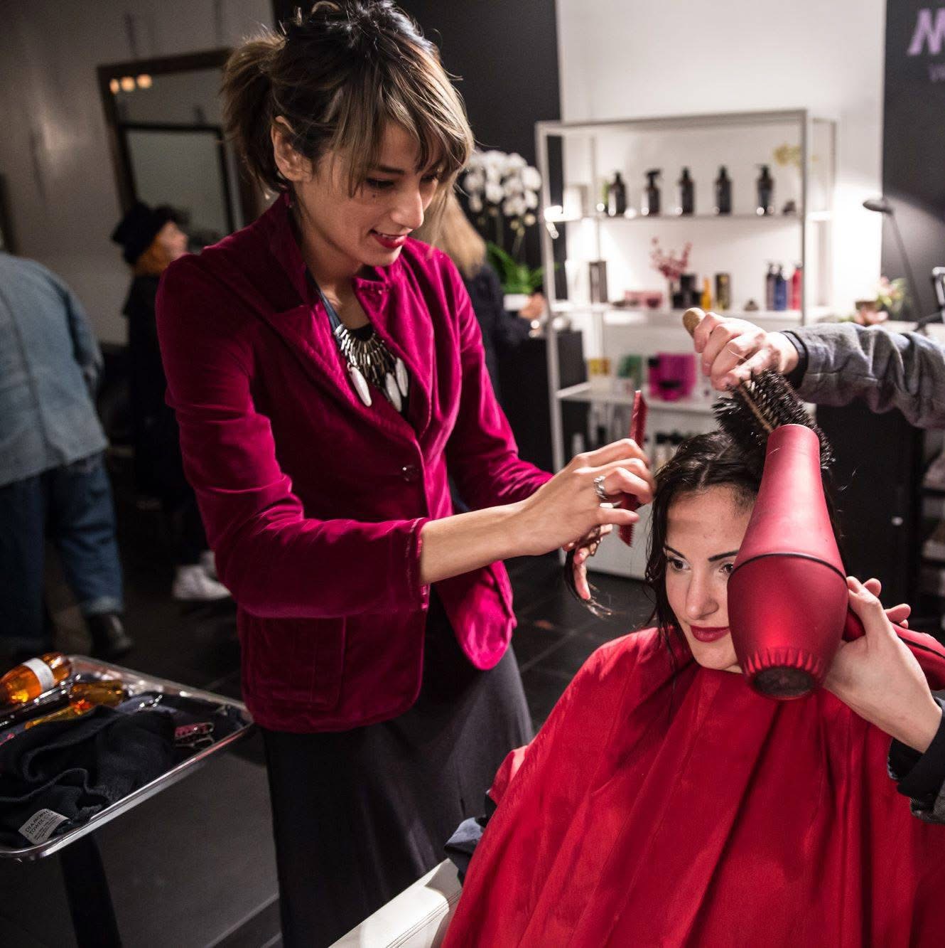 Woman getting her hair styled at a salon, with a stylist using a hairdryer, in a modern interior with shelves and products.