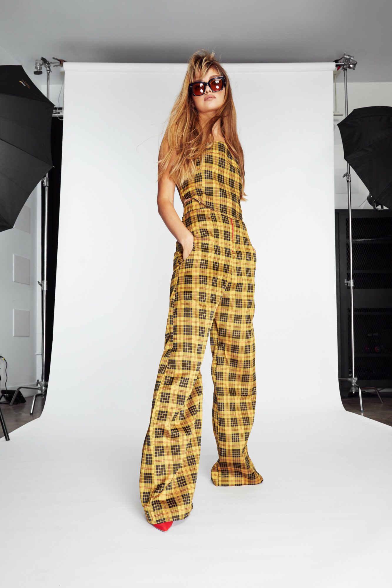 Woman with long hair wearing sunglasses and a yellow plaid jumpsuit, standing in a photography studio with a white backdrop.