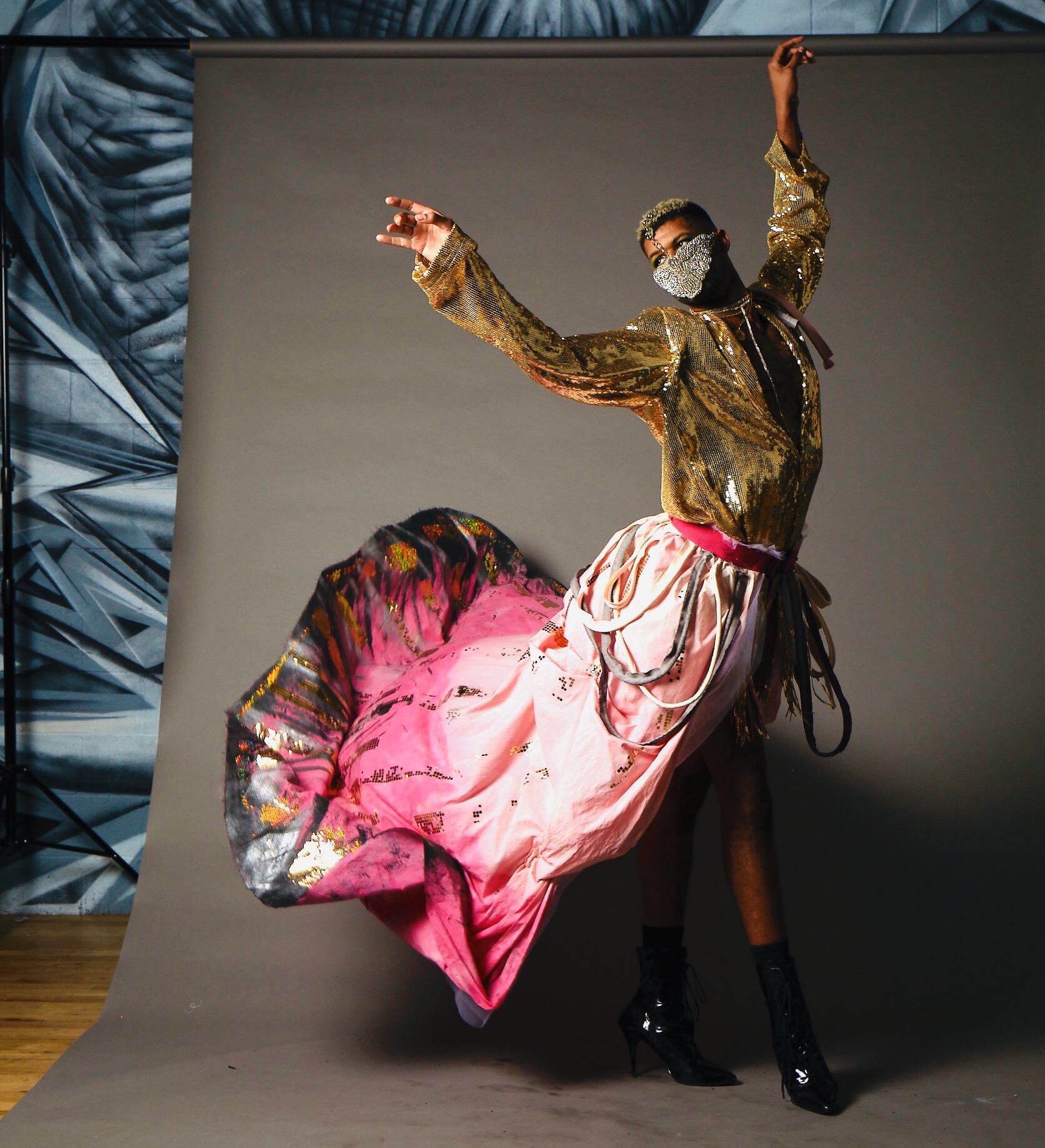 Person in a gold jacket, pink skirt, black boots, and face mask dancing with one arm raised, against a plain background.