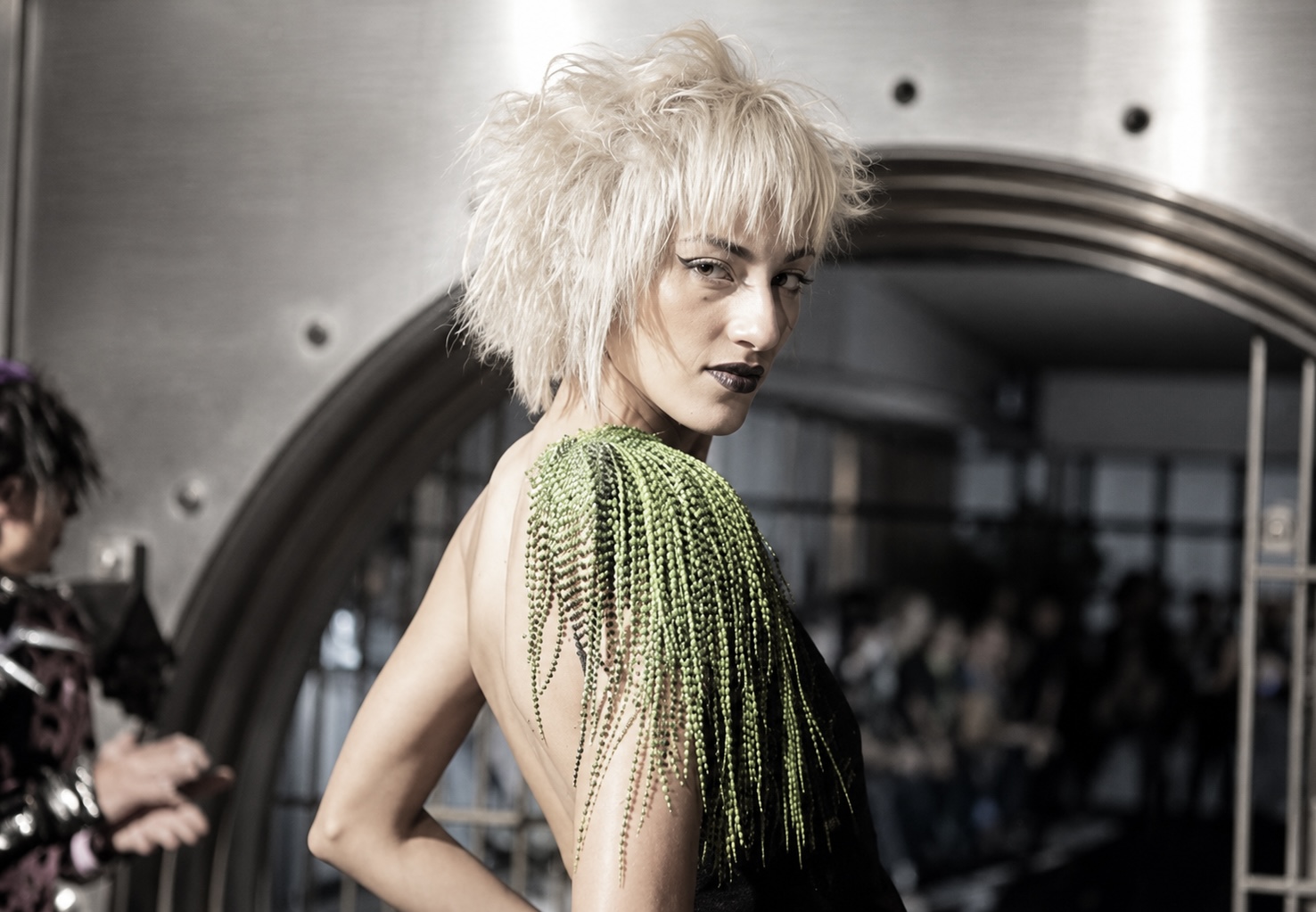 Person with short, curly, platinum blonde hair and dark makeup, wearing a green and black outfit, in an indoor setting.