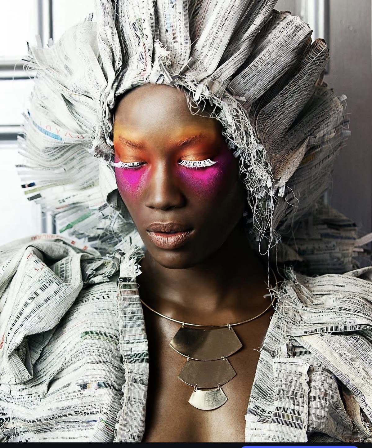 Person with newspaper headdress and clothing, vibrant pink eye makeup, and metallic necklace, standing near a window.