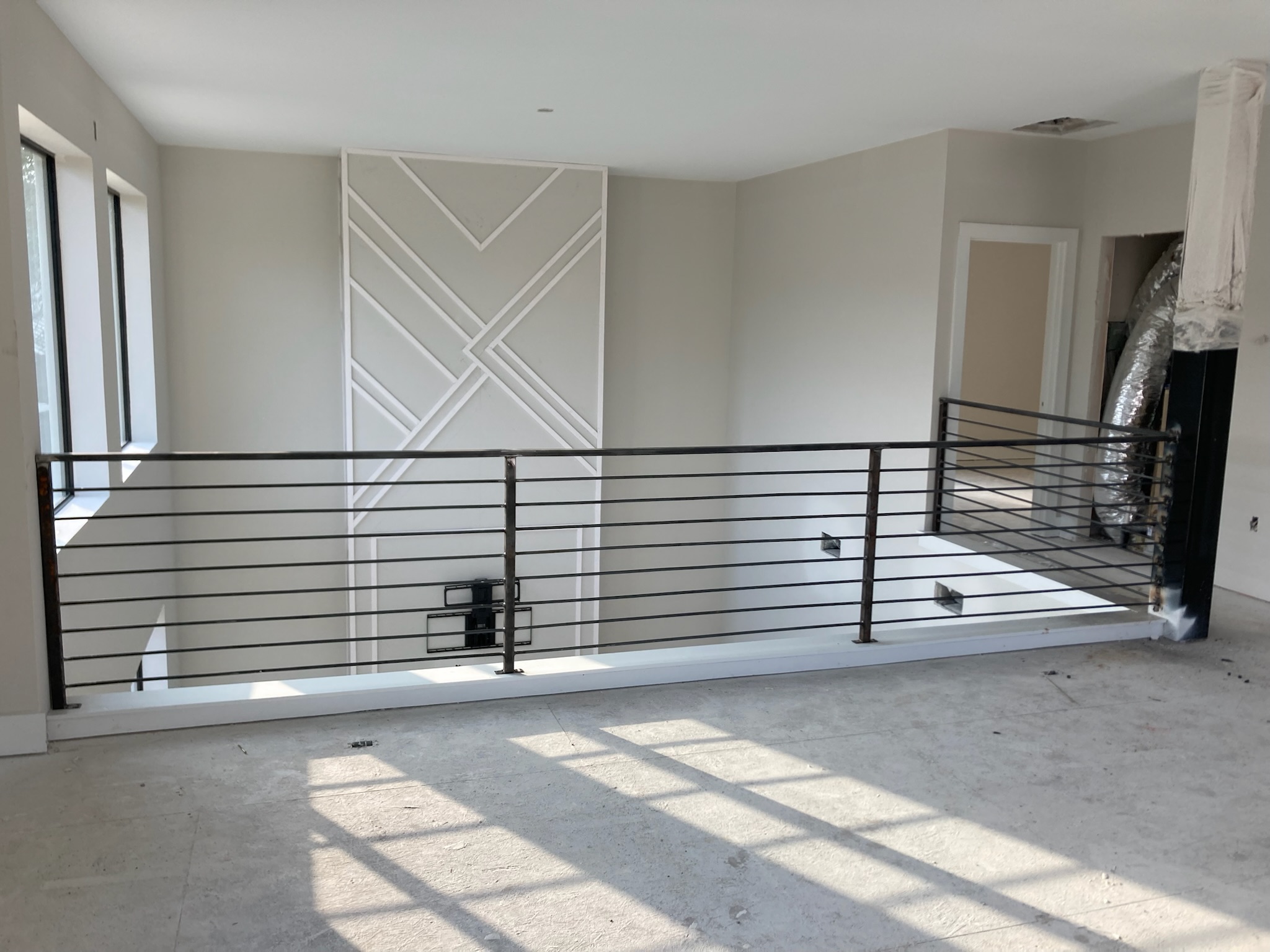 Interior space with a metal railing, large window, and decorative wall panel, sunlight casting shadows on the floor.