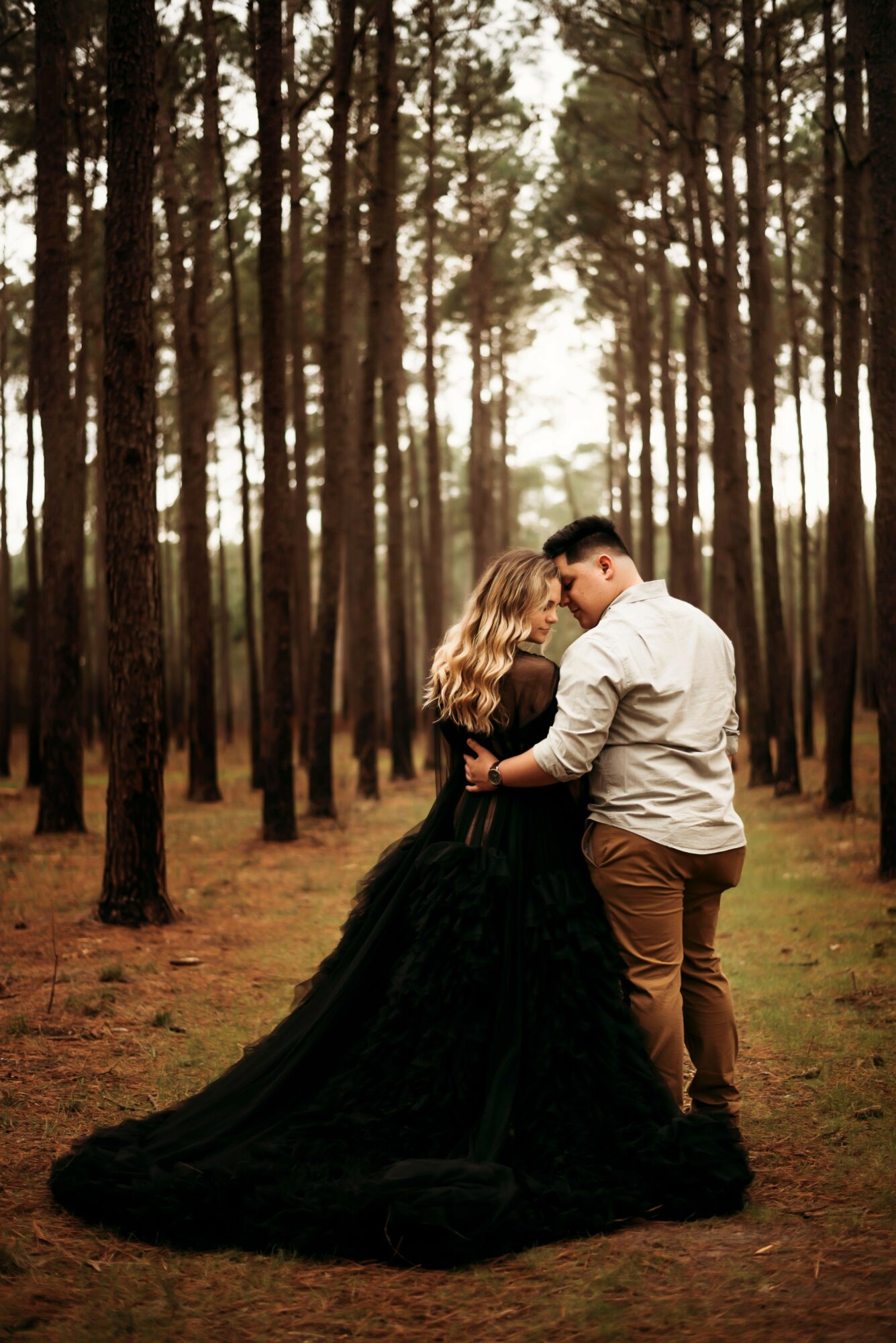 A couple dancing in a forest with tall trees, sunlight filtering through, woman in a black dress and man in a white shirt.