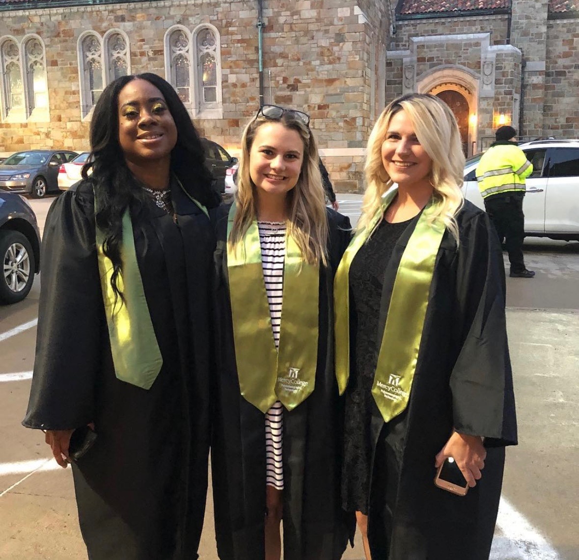 Three women in graduation gowns and stoles standing outdoors in front of a historic building, smiling.