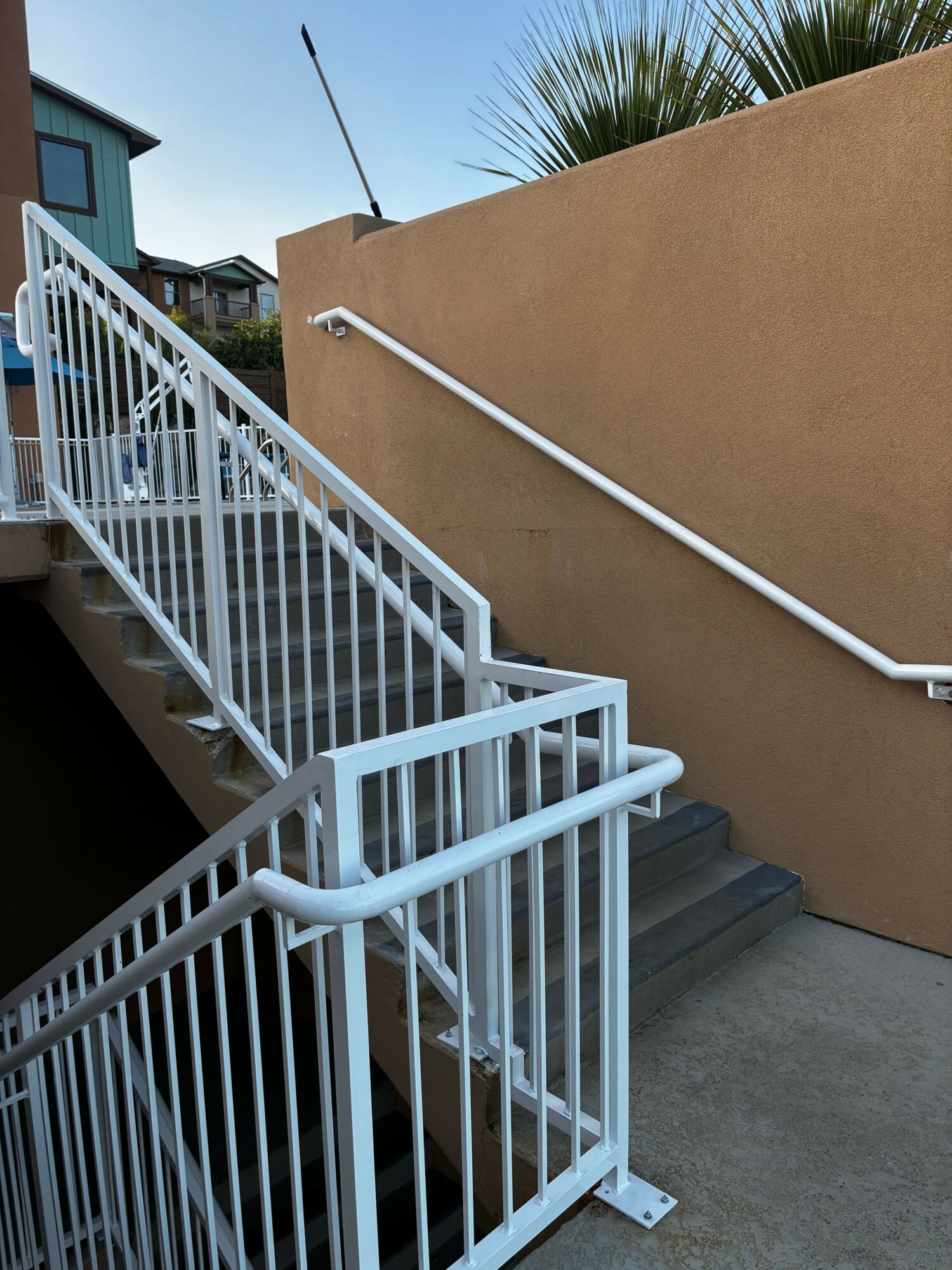 White metal staircase with railing outside a building, viewed from below, with stairs leading upward and a beige wall in background.