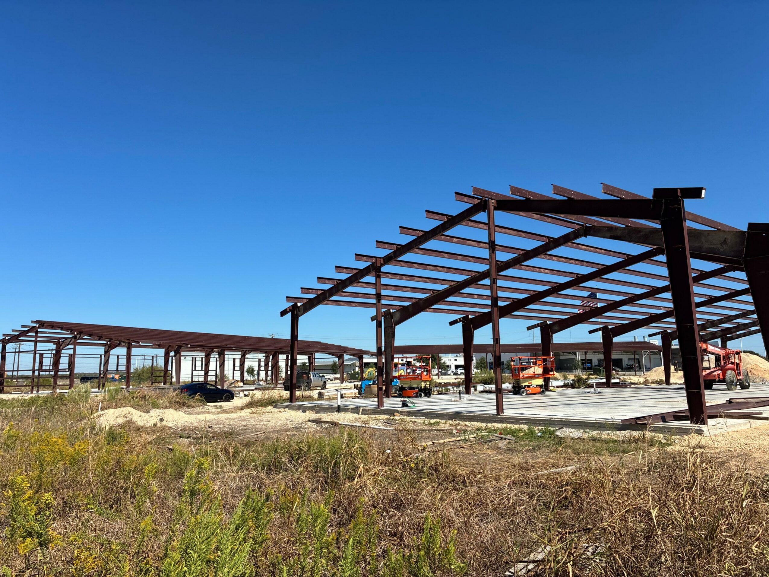 Construction site with steel framework under a clear blue sky, with grass and vegetation in the foreground.