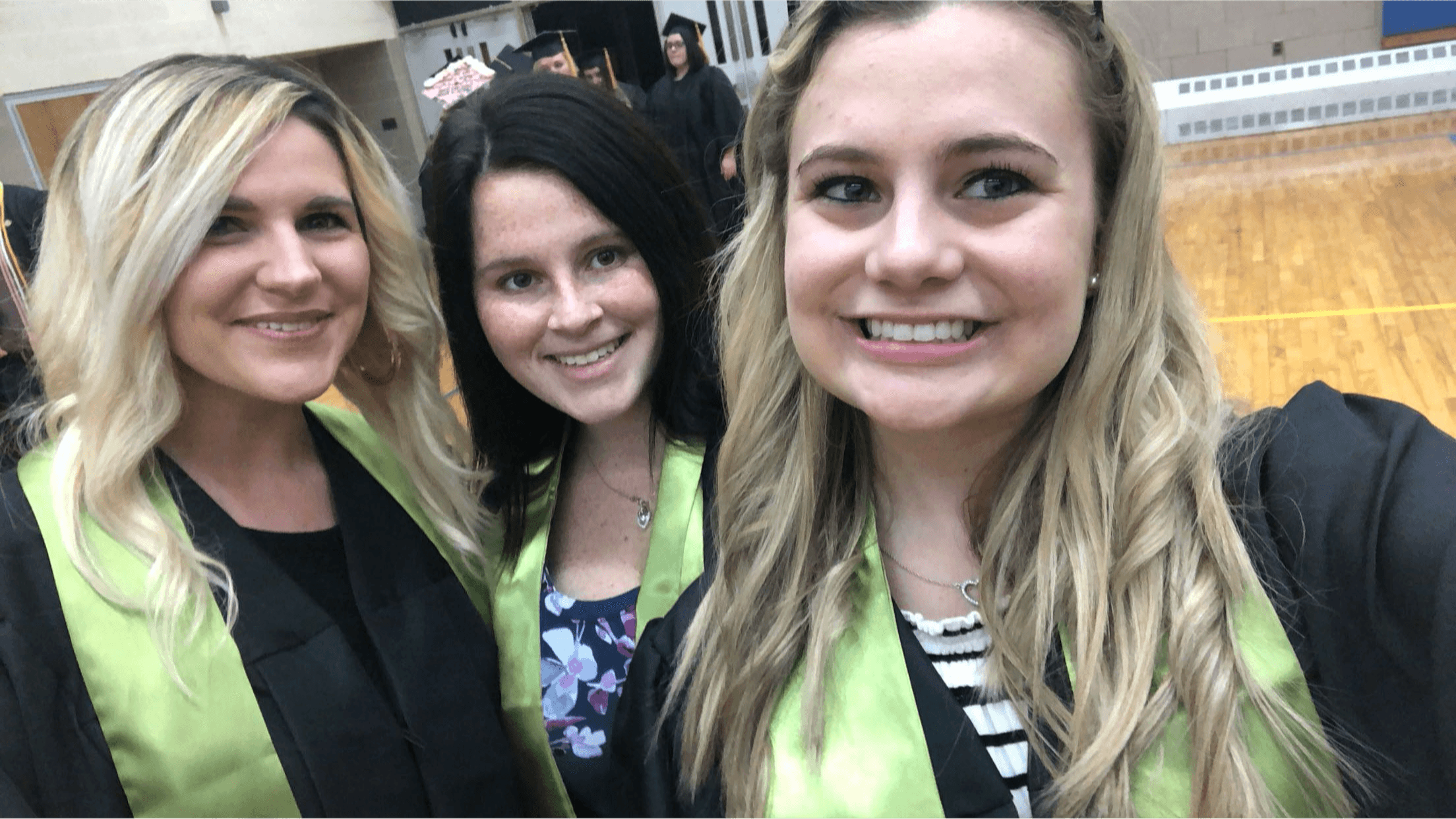 Three women smiling, standing indoors, two wearing lime green vests, one in a black jacket, background shows a gymnasium floor.