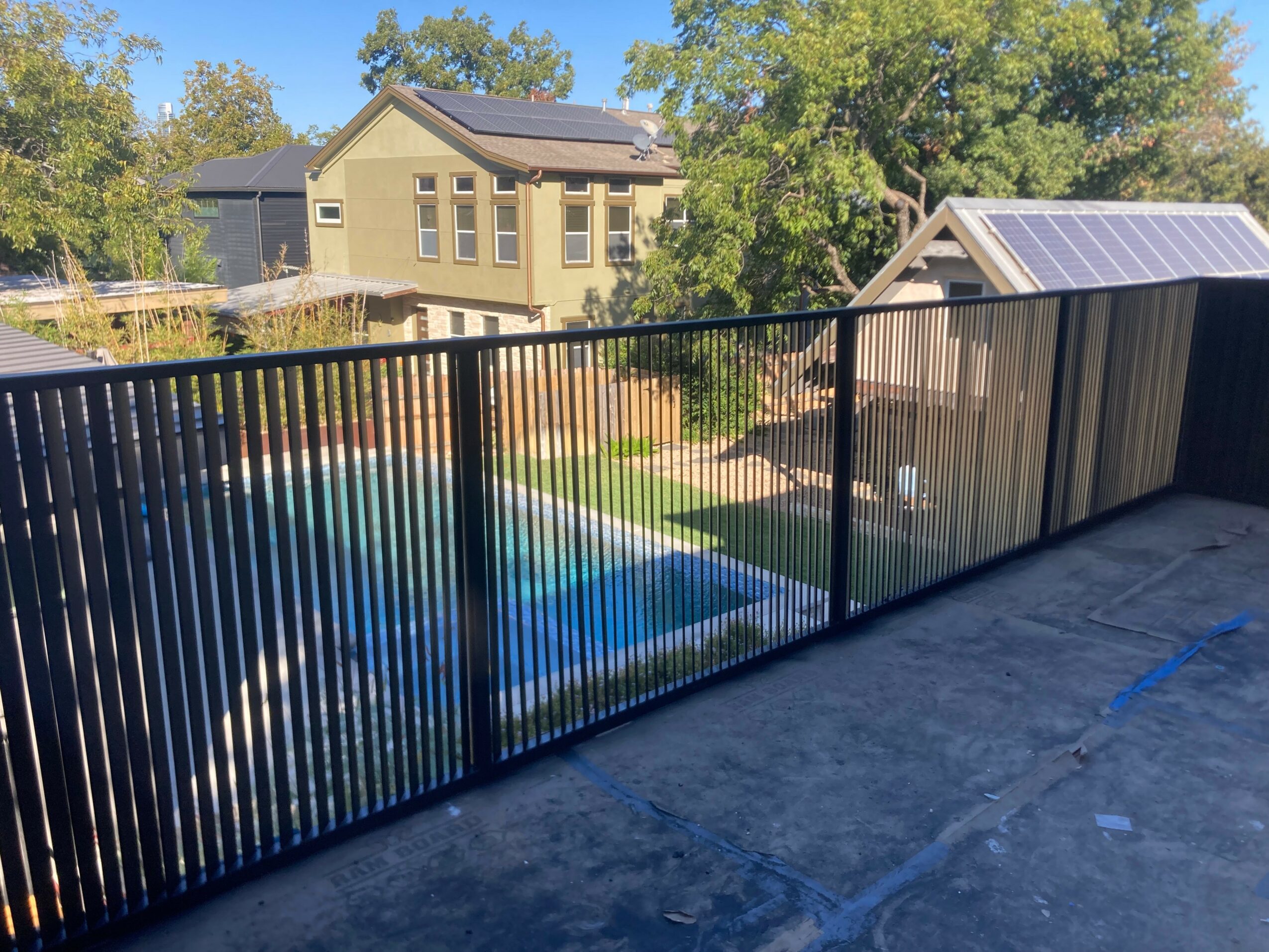 View from a balcony with a black metal railing overlooking a backyard with a swimming pool, trees, and houses in the background.