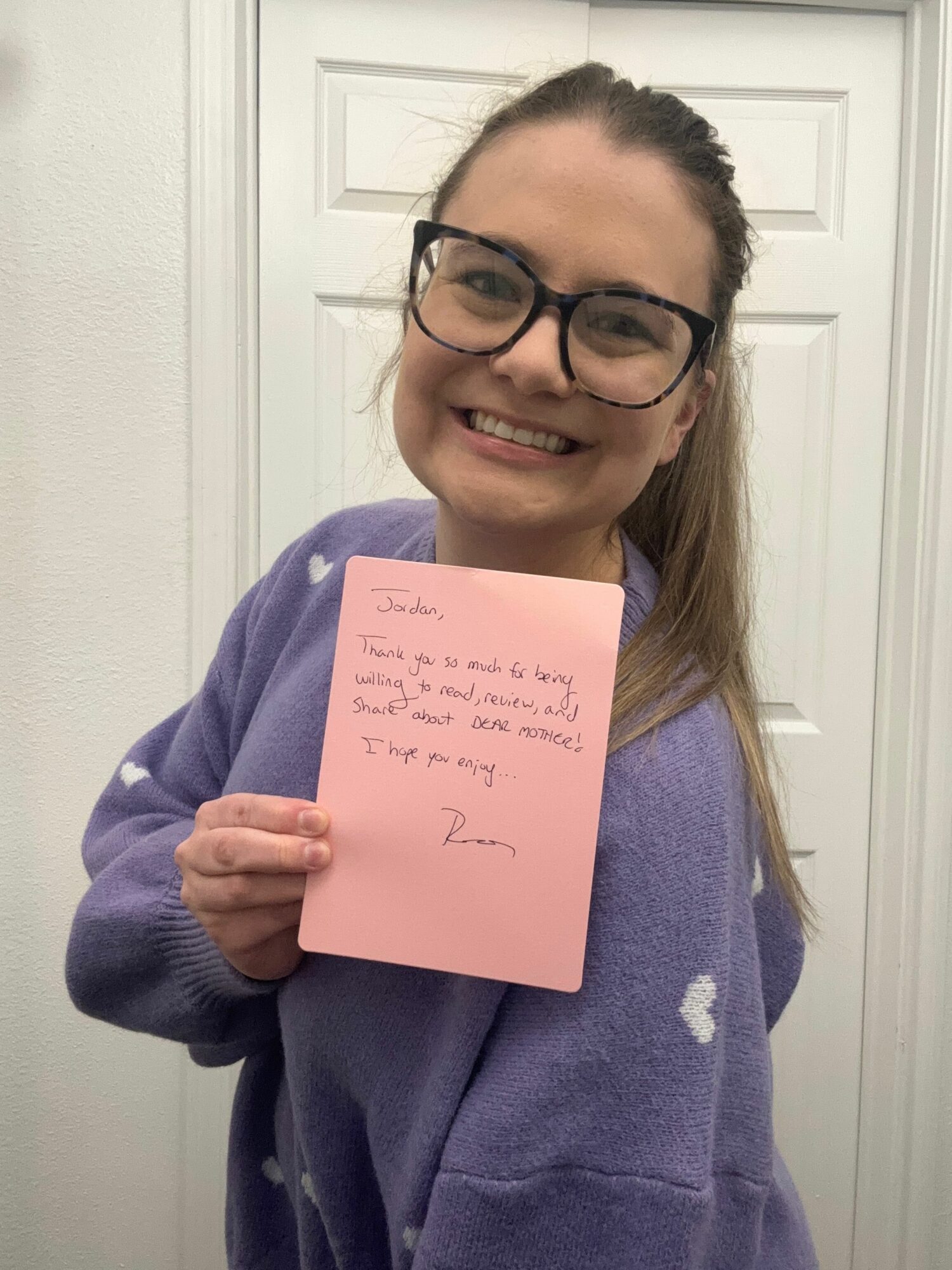 Smiling woman with glasses holding a pink note card, standing in front of a white door, wearing a purple hoodie.