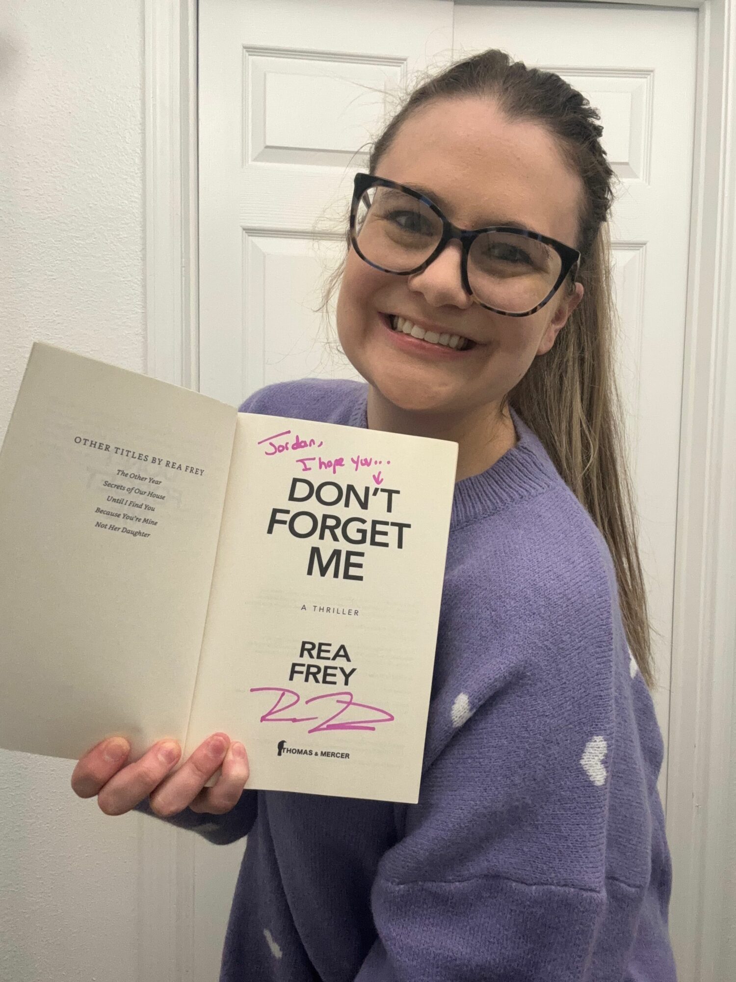 Woman with glasses smiling holding an open book, standing in front of a white door, wearing a purple sweater.