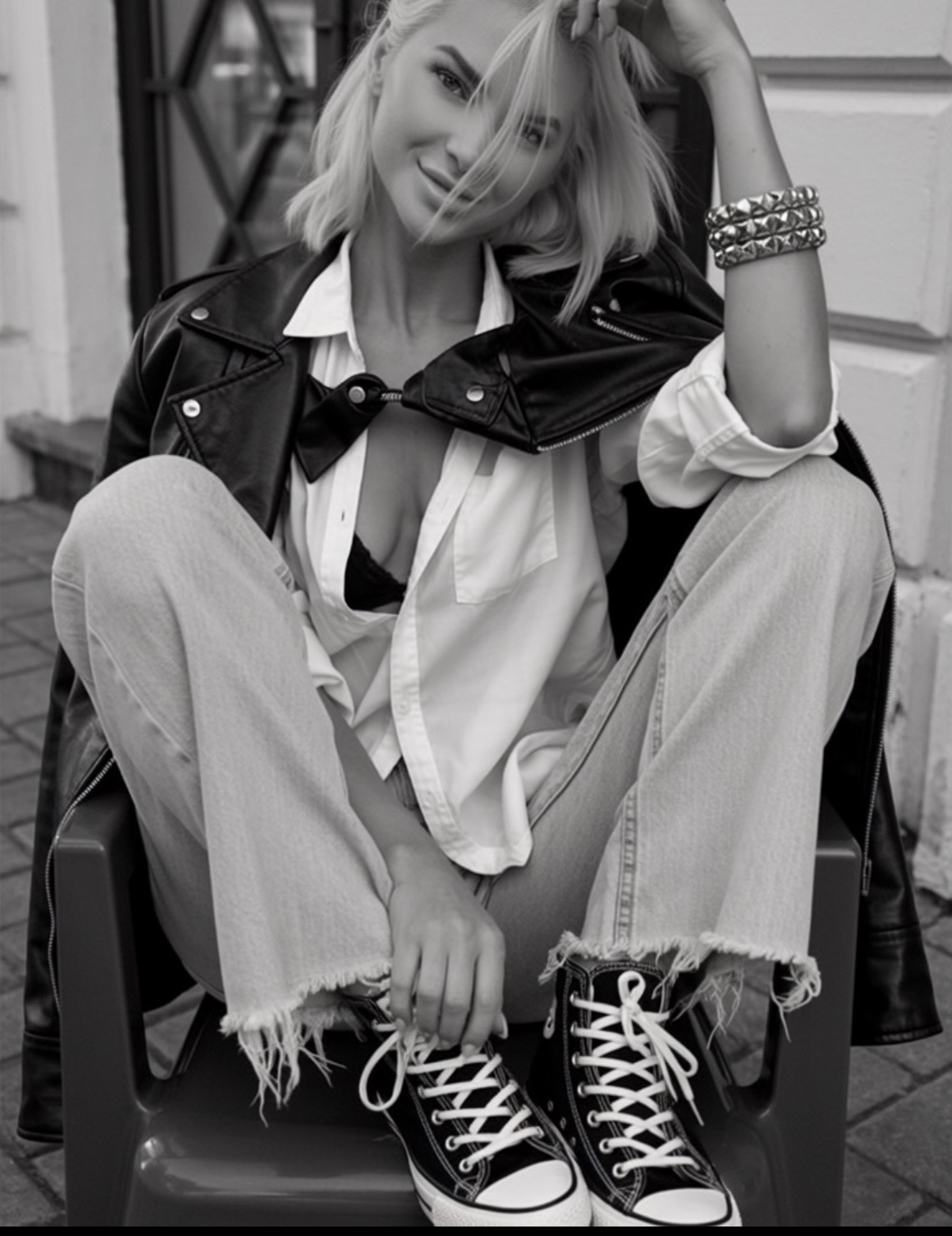 Woman sitting outdoors, smiling, wearing a leather jacket, loose shirt, ripped jeans, and sneakers, with her hand on her head.