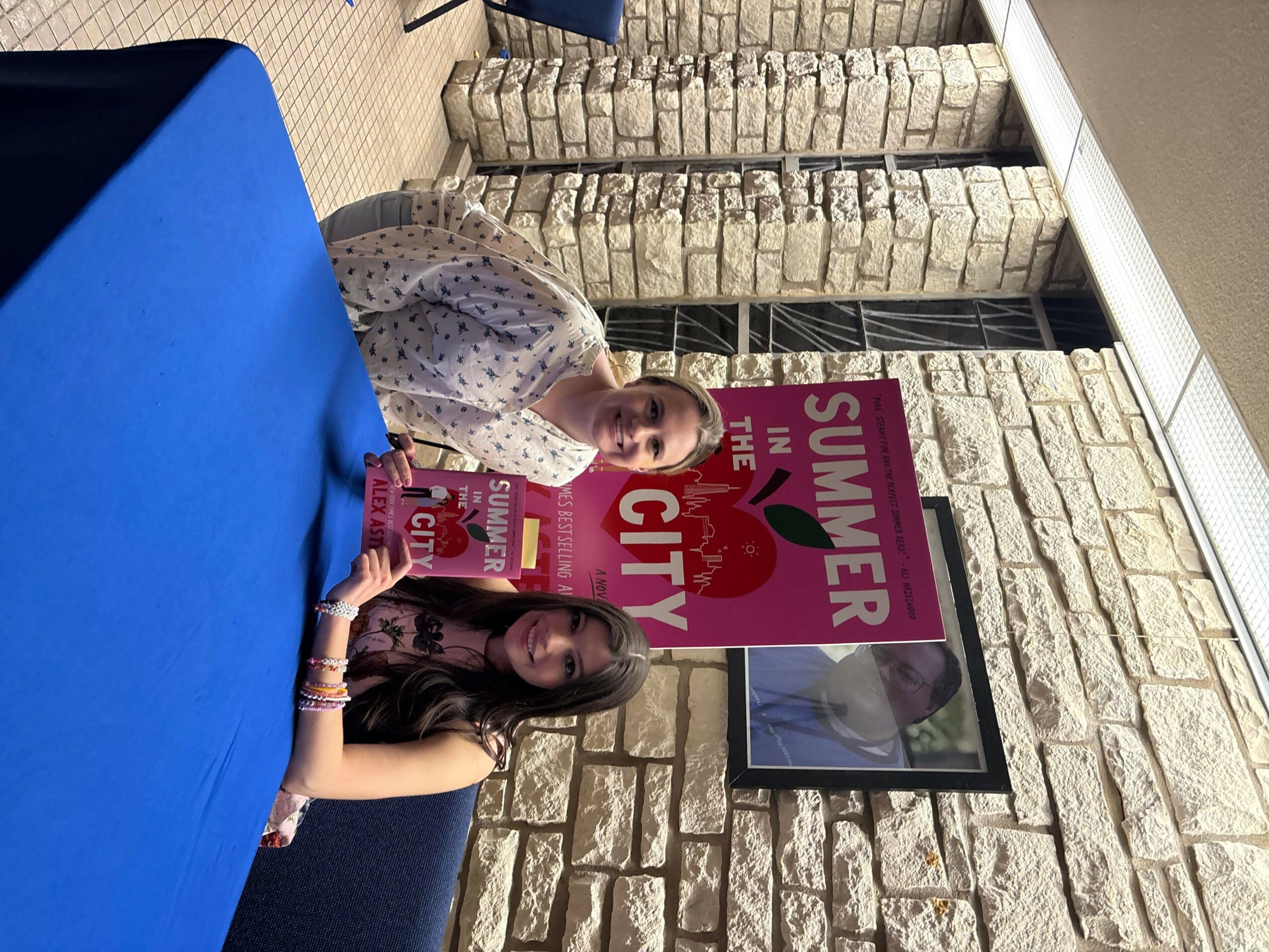 Two women sitting at a table with a blue tablecloth, holding a magazine, in front of a stone wall and a framed picture.