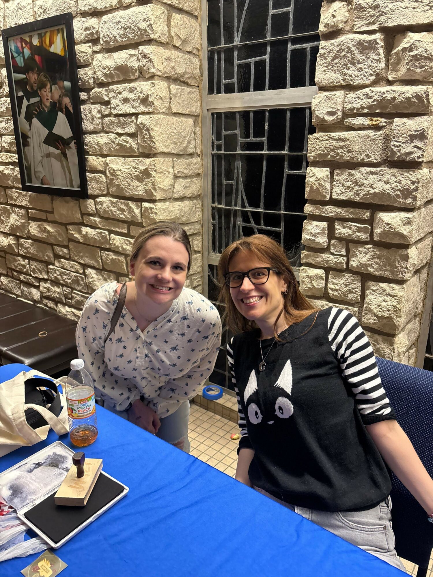 Two women sitting at a table with a stone wall background, smiling, with a tablet and a small trophy on the table.