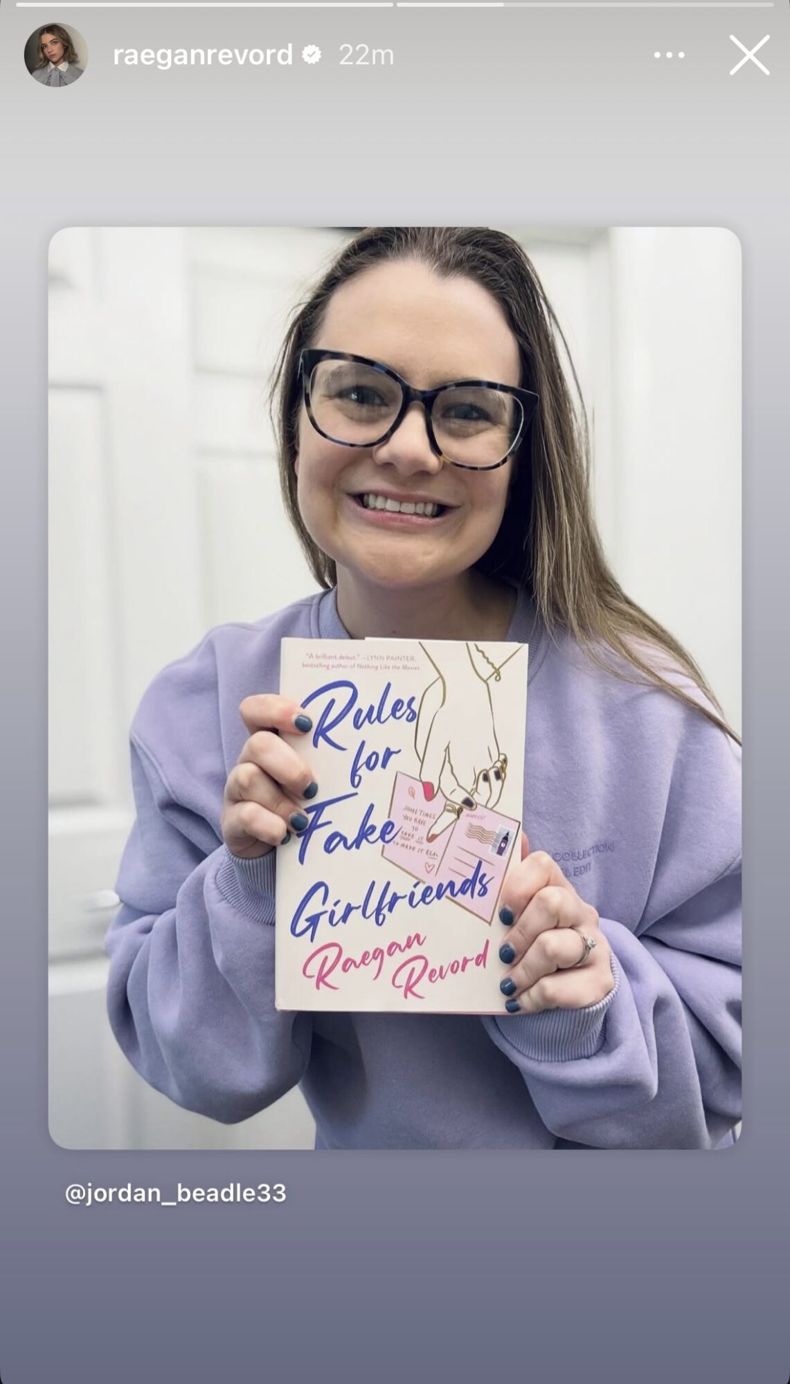 Young woman with glasses holding a book titled 'Rules for Fake Girlfriends' smiling, wearing a purple hoodie, indoors.