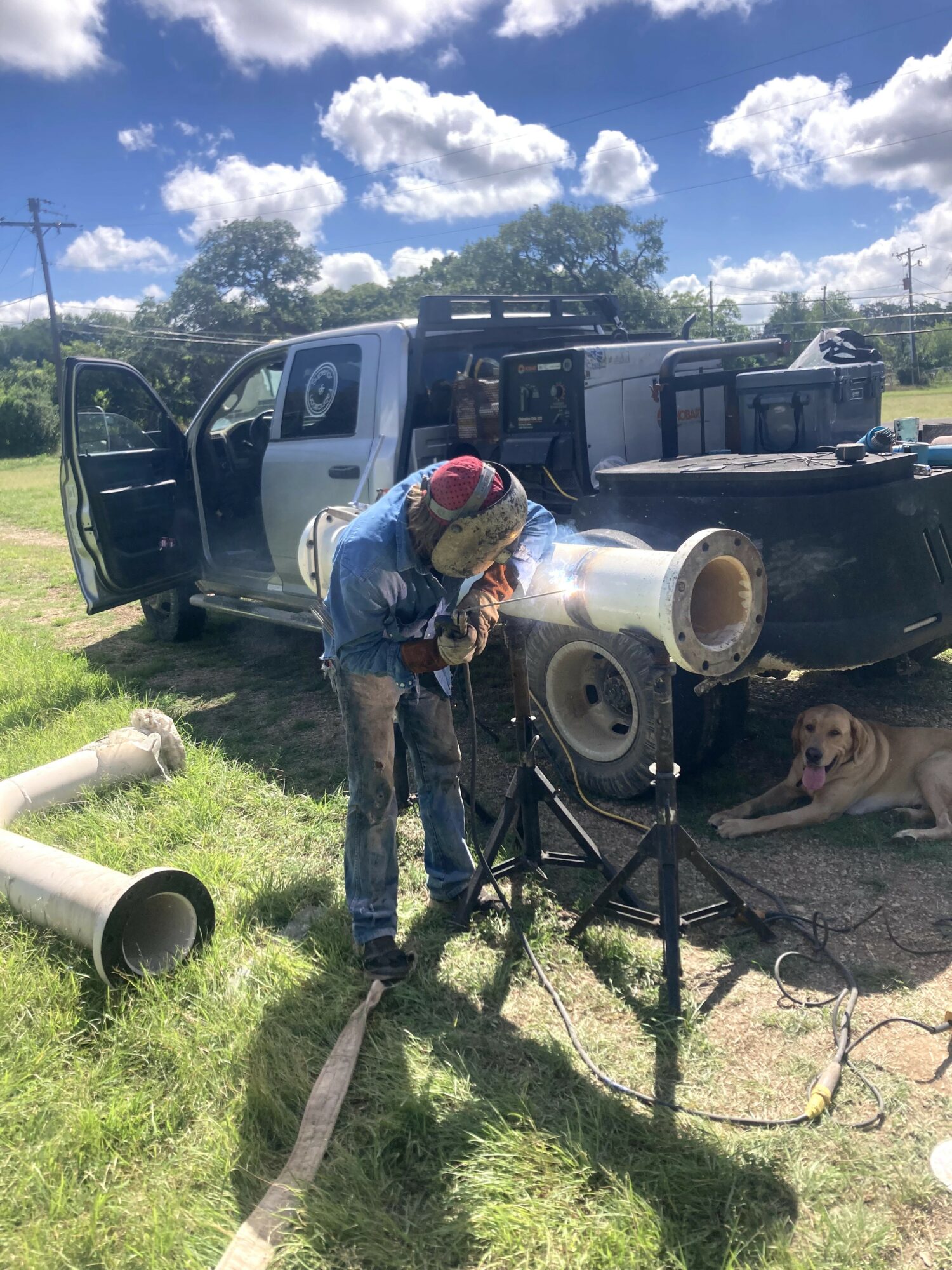 Person working with equipment near a pickup truck outdoors, with a dog lying nearby, under a partly cloudy sky.