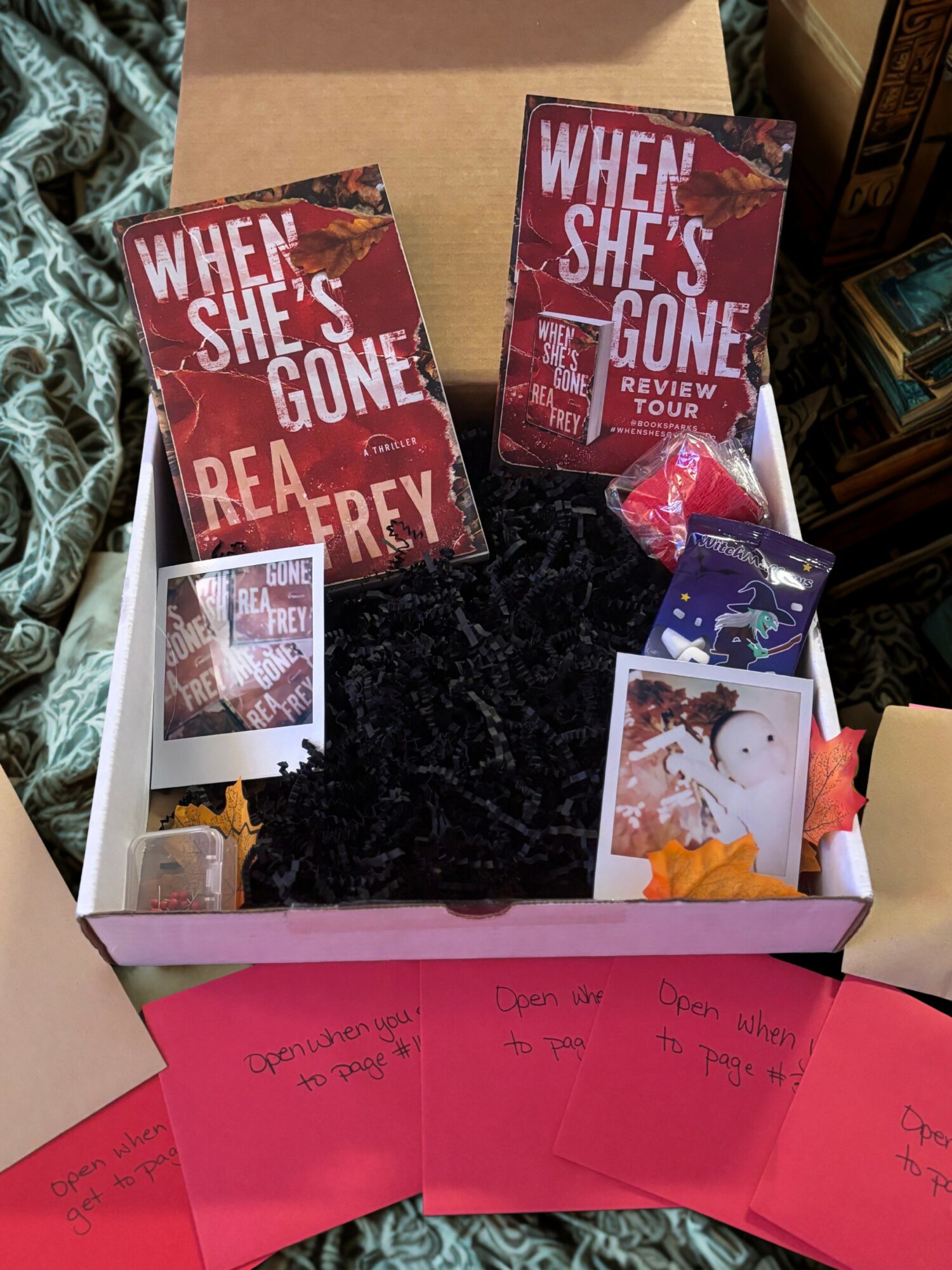 Open box with book and promotional materials, surrounded by pink notes and fabric, on a dark surface.