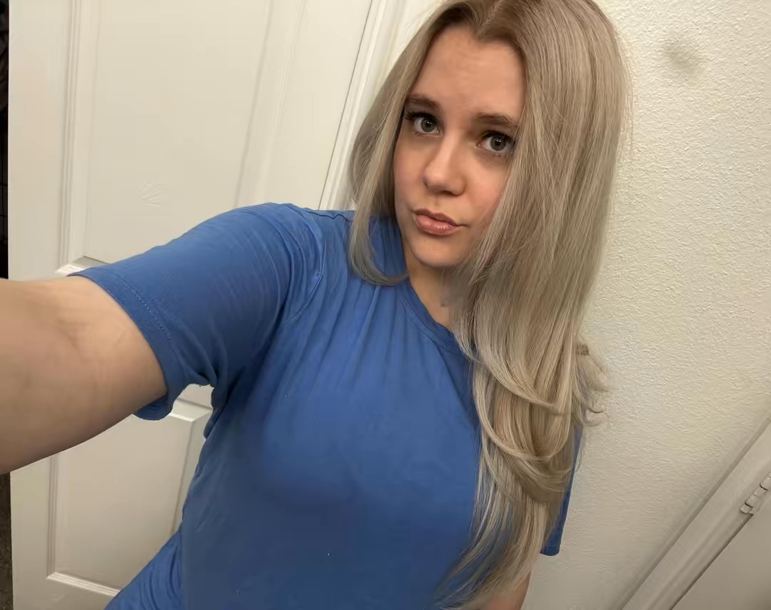 Young woman with long blonde hair wearing a blue shirt, taking a selfie indoors against a white wall.