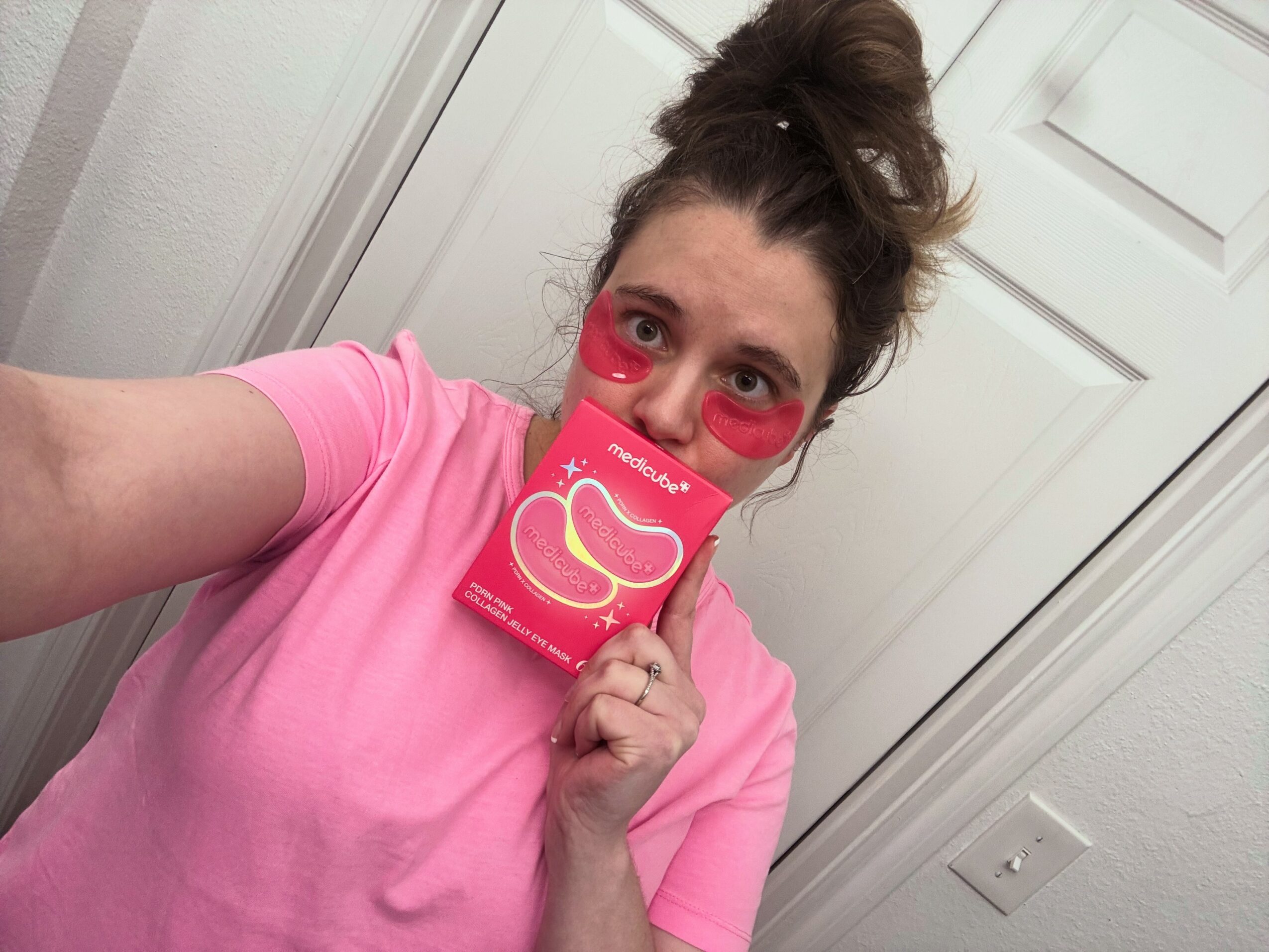 Person with pink face mask holding a pink box, wearing a pink shirt, standing in front of a white door.