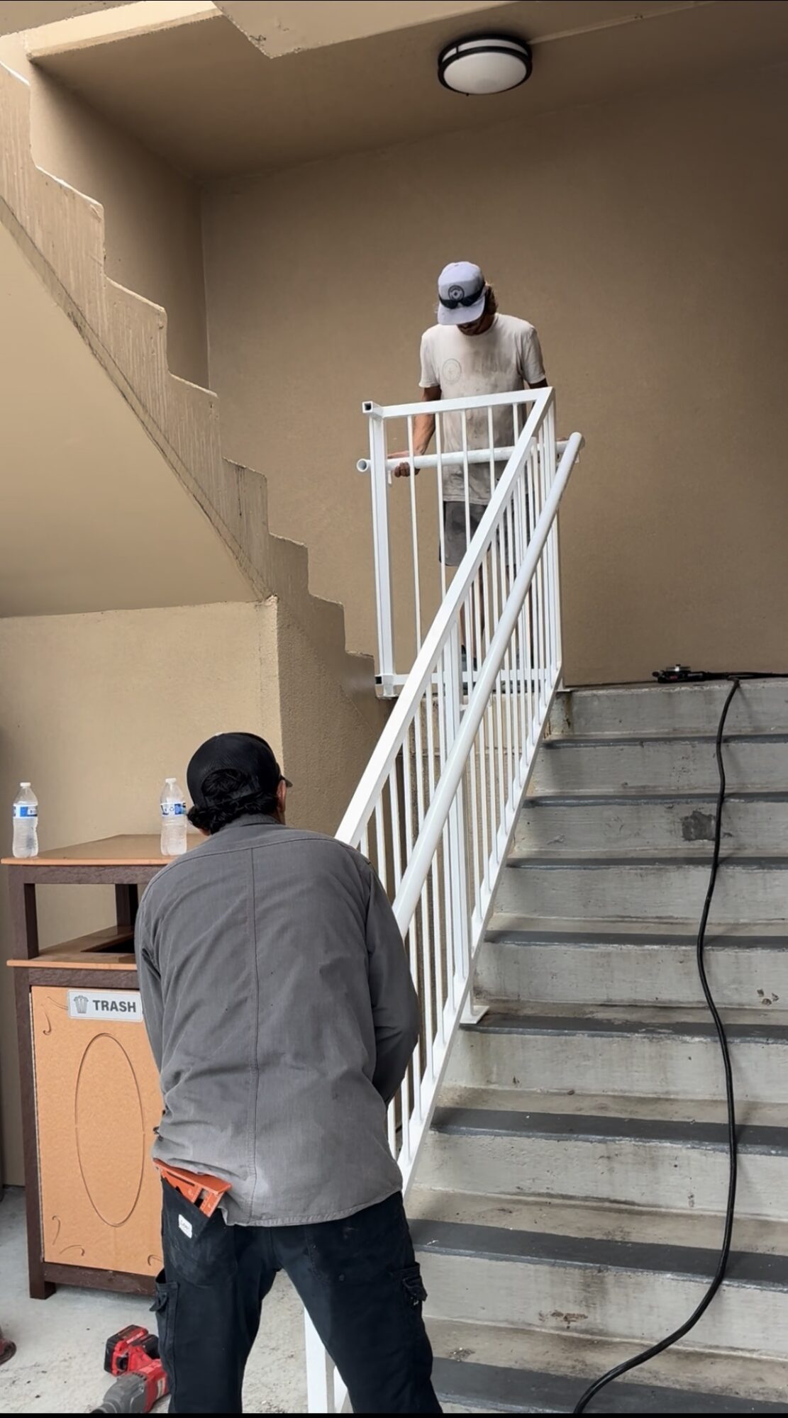 Person in gray jacket standing at bottom of stairs, person in white outfit and helmet on upper landing, beige wall background.