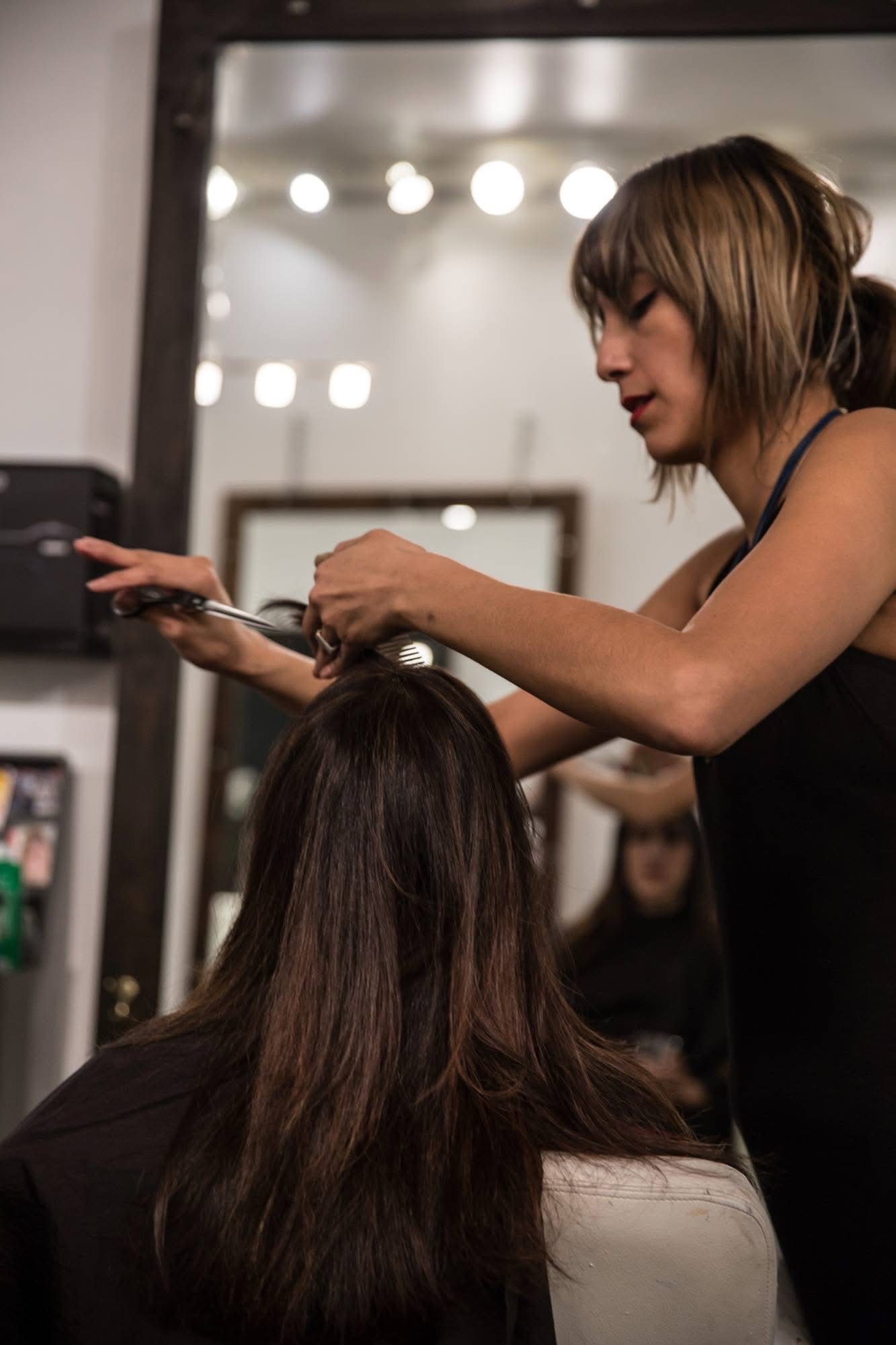 Hairdresser styles a woman's hair in a salon with bright lights and mirrors, focusing on her hair and hands.