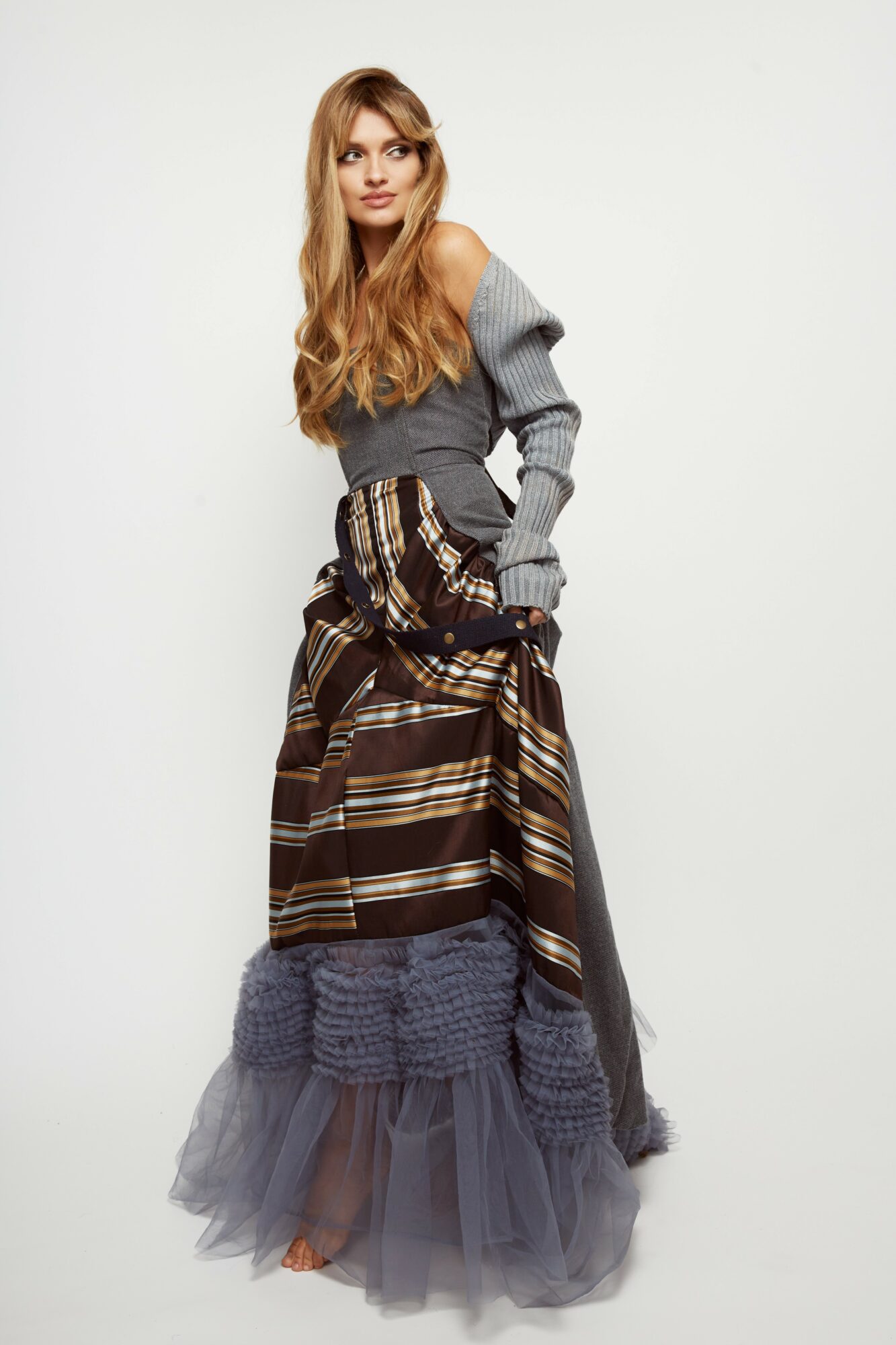 Woman with long hair wearing a gray off-shoulder sweater and layered skirt, standing against a plain white background.